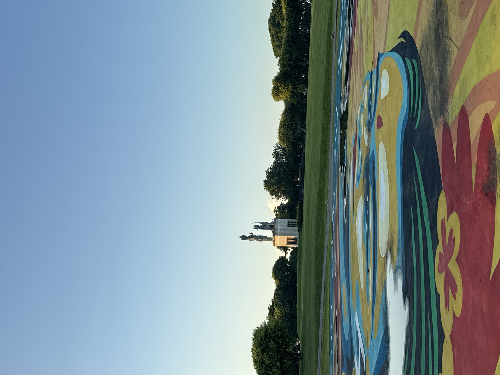 The image shows a view of a park with a colorful painted surface in the foreground. In the distance, a statue is visible. The sky is clear blue. The location is Kuta Selatan, Indonesia.