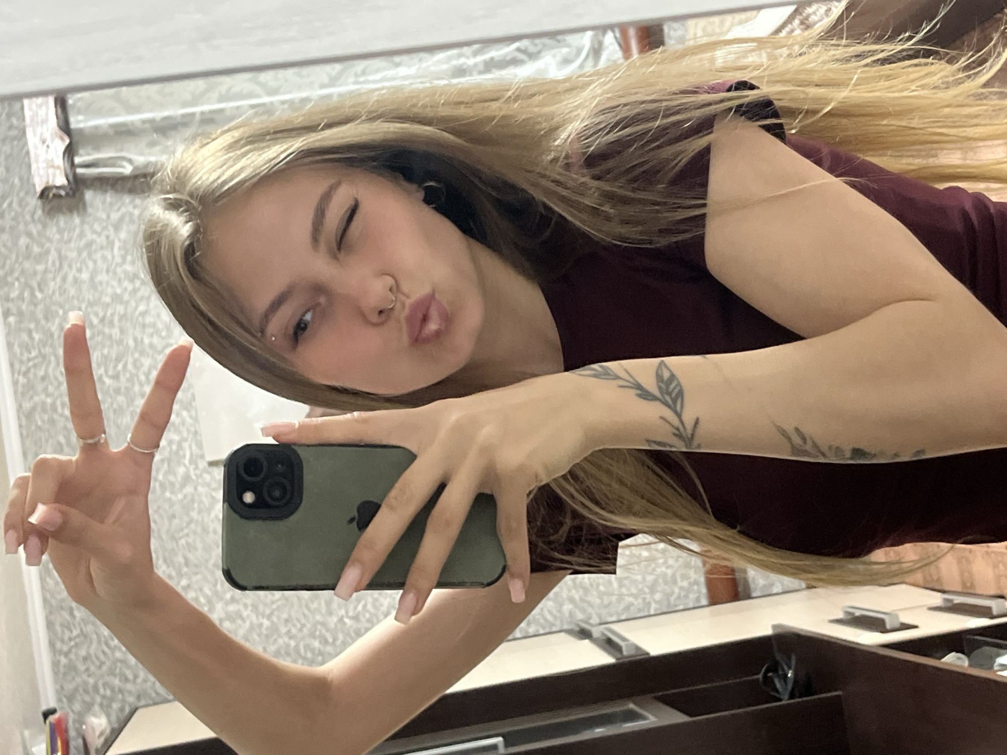 A young adult female is depicted in a mirror selfie taken indoors in the city of Фоки, Russia. She has long, light blonde hair and is wearing a dark maroon short-sleeved top. Her left hand is raised, forming a "peace" sign with two extended fingers, which are adorned with two silver-colored rings on the index and middle fingers. Her right hand holds a dark green smartphone with a visible Apple logo and a triple camera array, obscuring part of her face. She has a barbell piercing above her left eyebrow and a circular septum ring. Her facial expression includes a wink of her right eye and pursed lips. Tattoos of leafy branches are visible on her right forearm and bicep. The background, reflected in the mirror, features a light-colored, patterned wallpaper, a dark brown shelving unit on the left, and a lighter cabinet with silver handles behind her. A patterned curtain is visible to the right of the subject.