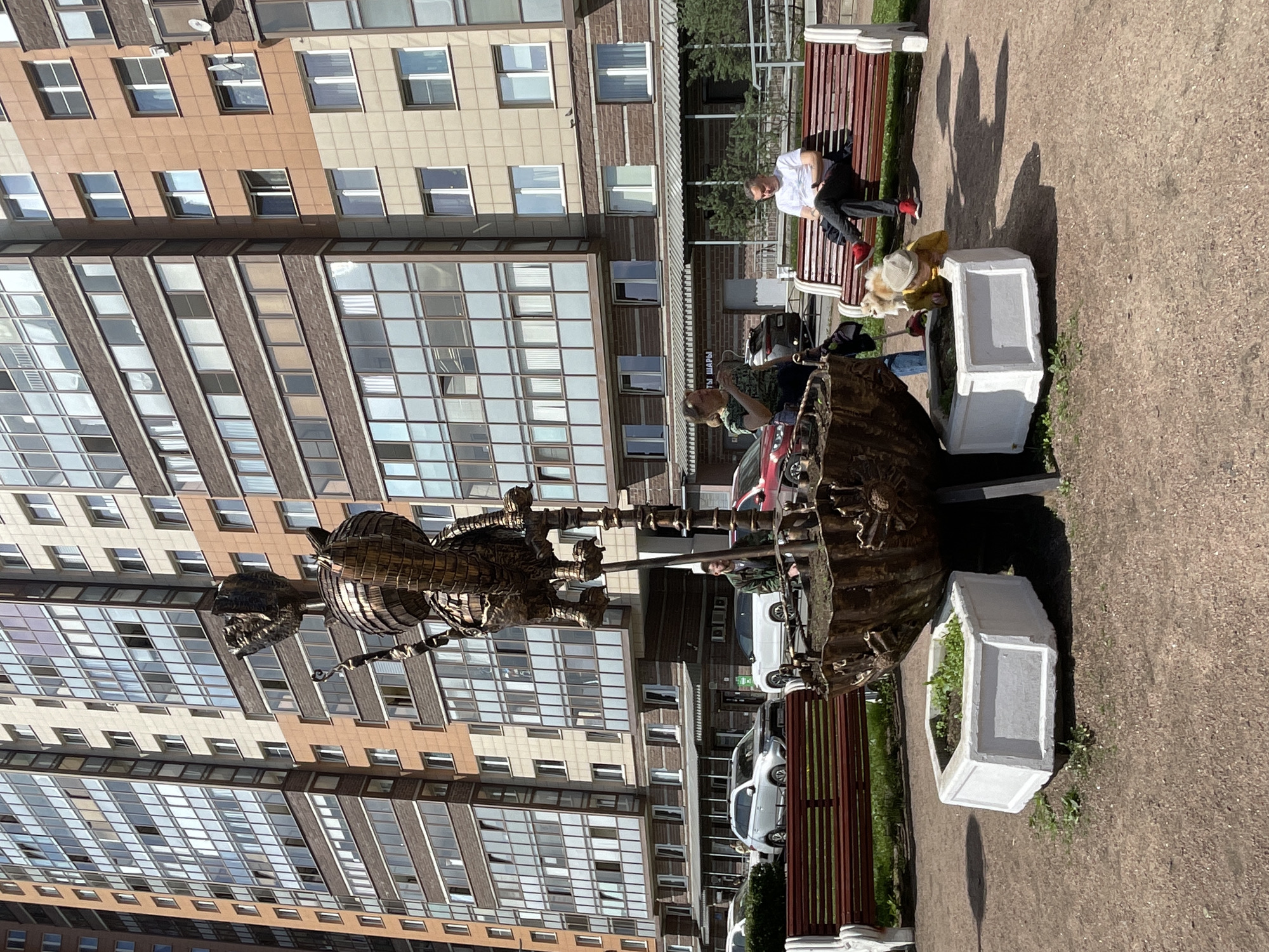 A daytime outdoor shot shows an artistic metal sculpture with figures in a public square in Saint Petersburg, Russia. The sculpture sits on a concrete platform. A person wearing a green shirt looks down at the sculpture, standing next to a red car. A bench is nearby, where a man in a white shirt and dark pants sits. A woman with a light-colored coat and a dog are seated near the bench. In the background, a modern high-rise apartment building with many windows rises. Several cars are parked behind the sculpture. The ground is covered in brown gravel. White concrete planters border the area.