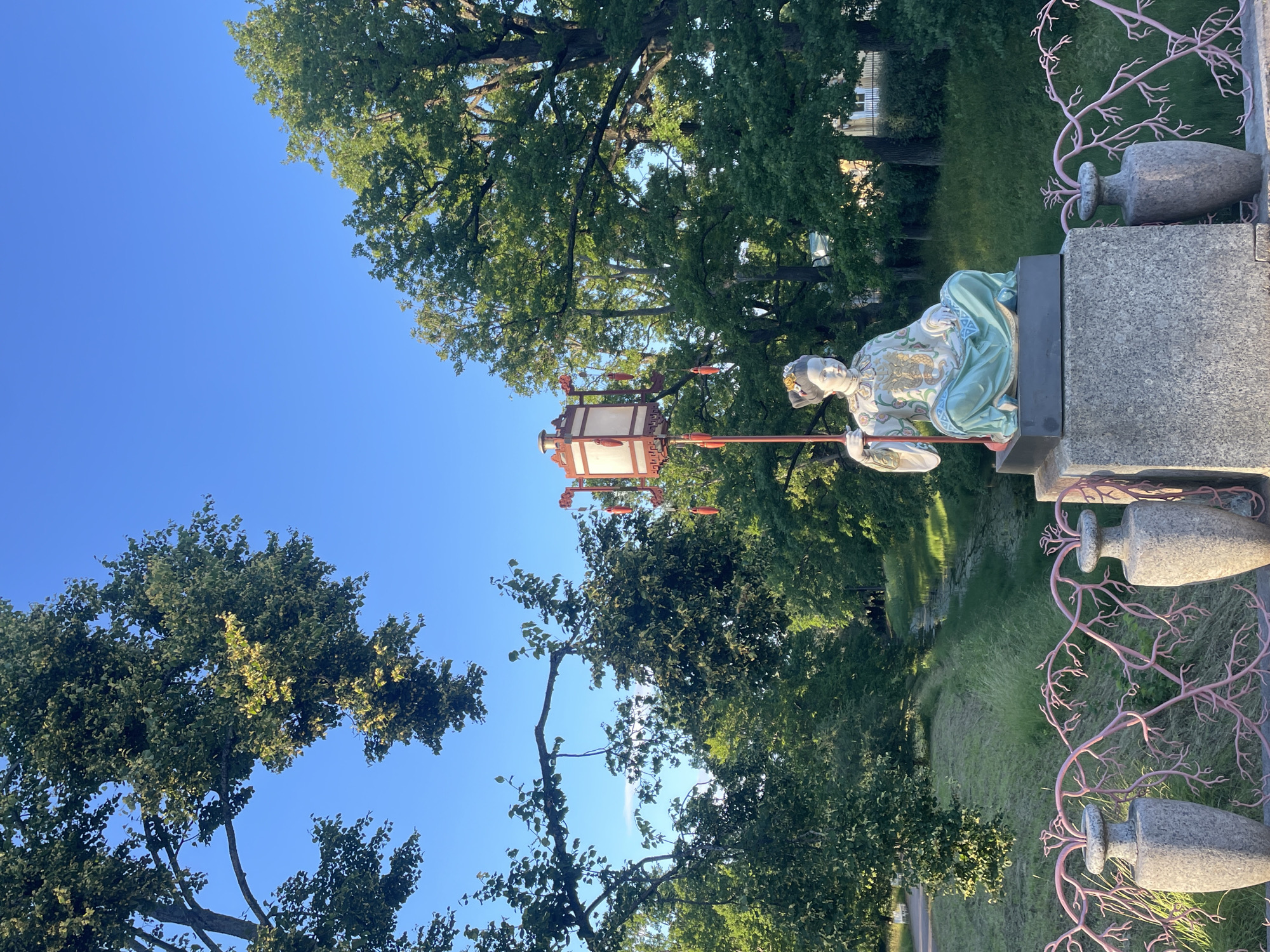 The image shows a statue of a seated figure holding a lantern in a park. The statue is on a pedestal made of stone. The figure is dressed in traditional Chinese clothing, with elaborate patterns on the robe and a headdress adorned with flowers. The lantern is rectangular with a decorative frame and tassels hanging from it. In the background, there are numerous trees with lush green foliage, and a clear blue sky. A narrow body of water, possibly a canal or stream, runs through the grassy area behind the statue. The foreground features a decorative pink railing with vase-like structures on either side. This statue is located in the Chinese Garden of Catherine Park, in the city of Pushkin, Russia, which is part of the Tsarskoye Selo museum-reserve.