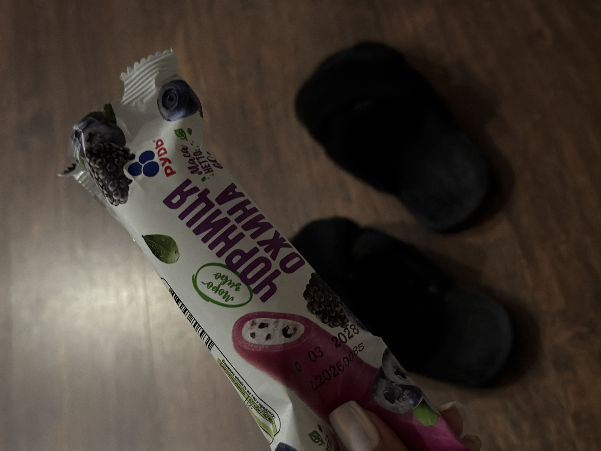 An indoor, close-up, eye-level shot shows a white ice cream wrapper with purple text and images of berries. The wrapper is diagonally positioned from the top left to the bottom right. The text on the wrapper includes "РУДЬ", "ЧОРНИЦЯ", and "ОЖИНА". Some numerical and text details are visible, including a date stamp "10 03 2028" and "420260085". In the bottom right corner, two dark, out-of-focus objects, possibly shoes, are visible. A person's hand with a pale fingernail is holding the wrapper from the left side of the image. The background is a wooden surface.