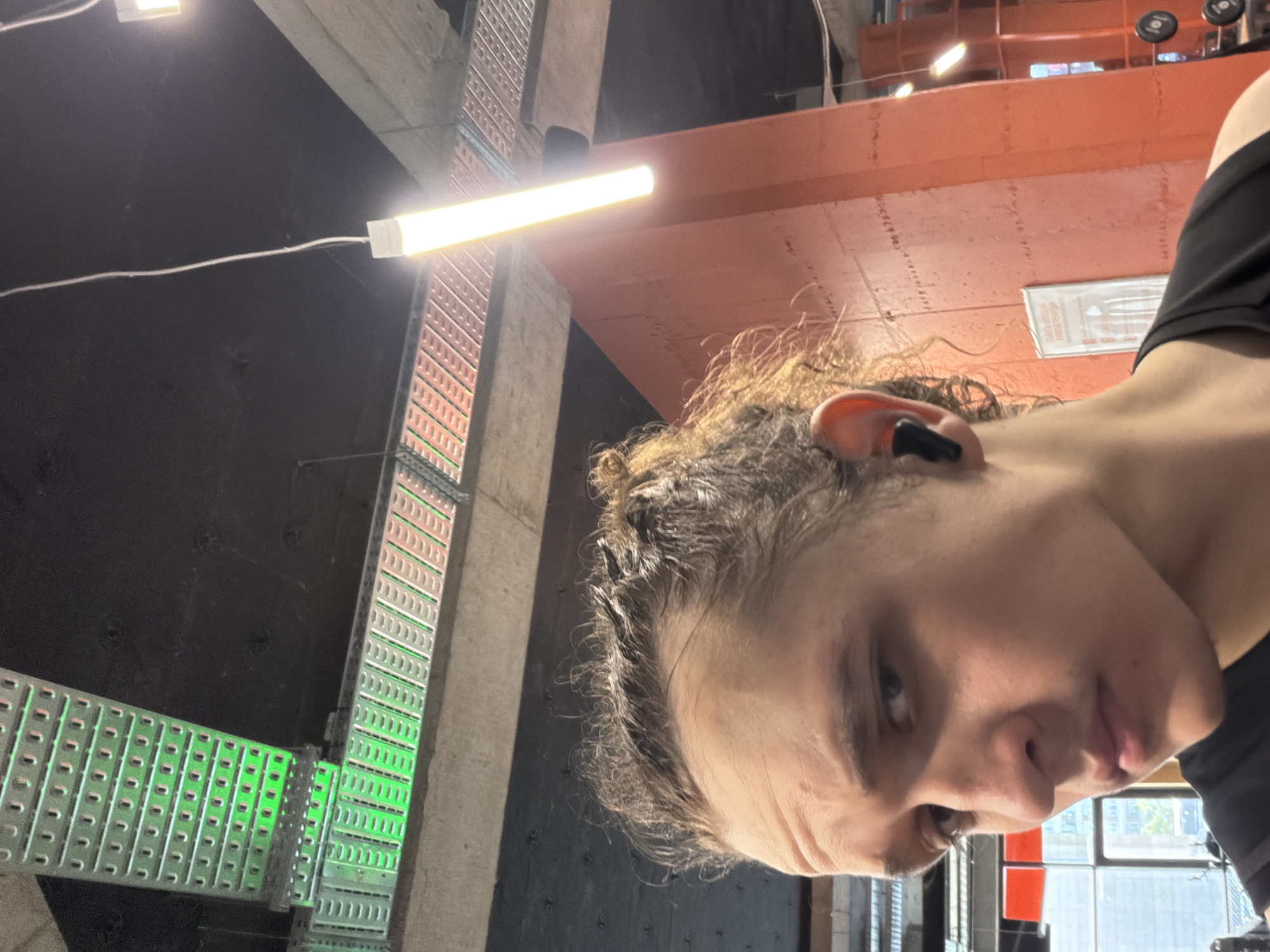 A low-angle, close-up selfie shot shows a person with light skin, short curly hair, and an earbud in their right ear. They are wearing a black top. The background reveals an industrial-looking gym environment. An overhead light fixture is visible in the upper left. Further in the background are concrete supports, metal cable trays (some illuminated in green), and red ceiling and wall elements. Dumbbells are partially visible at the top of the image. Through a window in the far background, buildings and sky can be seen. The precise location in Plovdiv, Bulgaria, cannot be determined from the visual information provided.