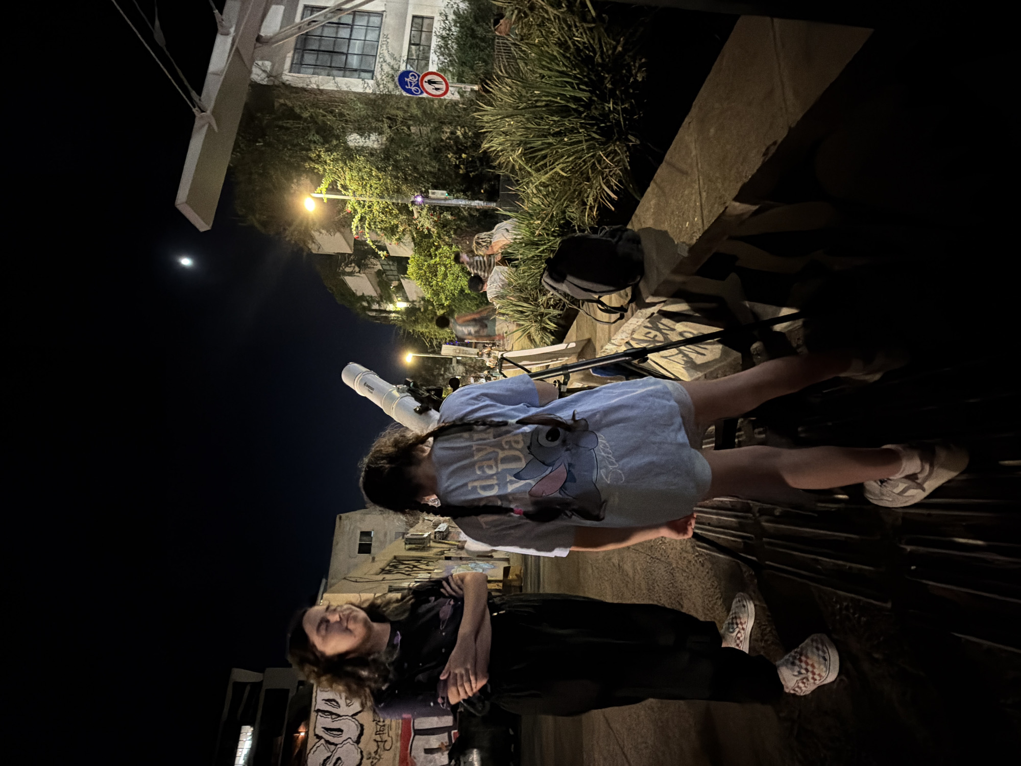 In Tel-Aviv, Israel, two girls are looking at the moon through a telescope at night. The girl on the left is wearing a black shirt with a purple pattern and black pants, with checkered Vans shoes. The girl on the right has her back to the camera, wearing a light blue t-shirt with a Stitch graphic and white sneakers. They are standing on a sidewalk in front of buildings with graffiti. The moon is visible in the dark sky. In the background, there are other people, trees, and a street lamp.