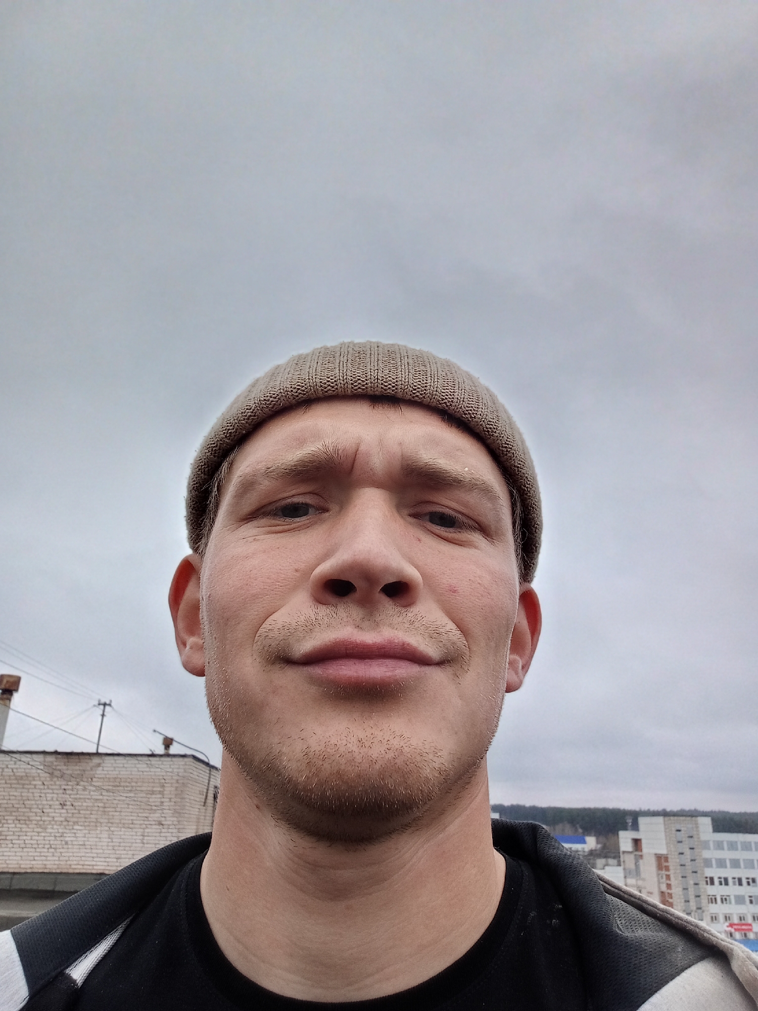 A close-up, low-angle selfie of a white male with a neutral expression. He is wearing a light brown beanie. His facial features include light-colored hair stubble, slightly furrowed brow, and a faint smile. He is wearing a black shirt with a white stripe along the shoulder. The background shows a cloudy sky and a cityscape, partially obscured by a brick building to the left. Buildings are visible in the distance. Based on known imagery, the location appears to be Chaikovsky, Russia.