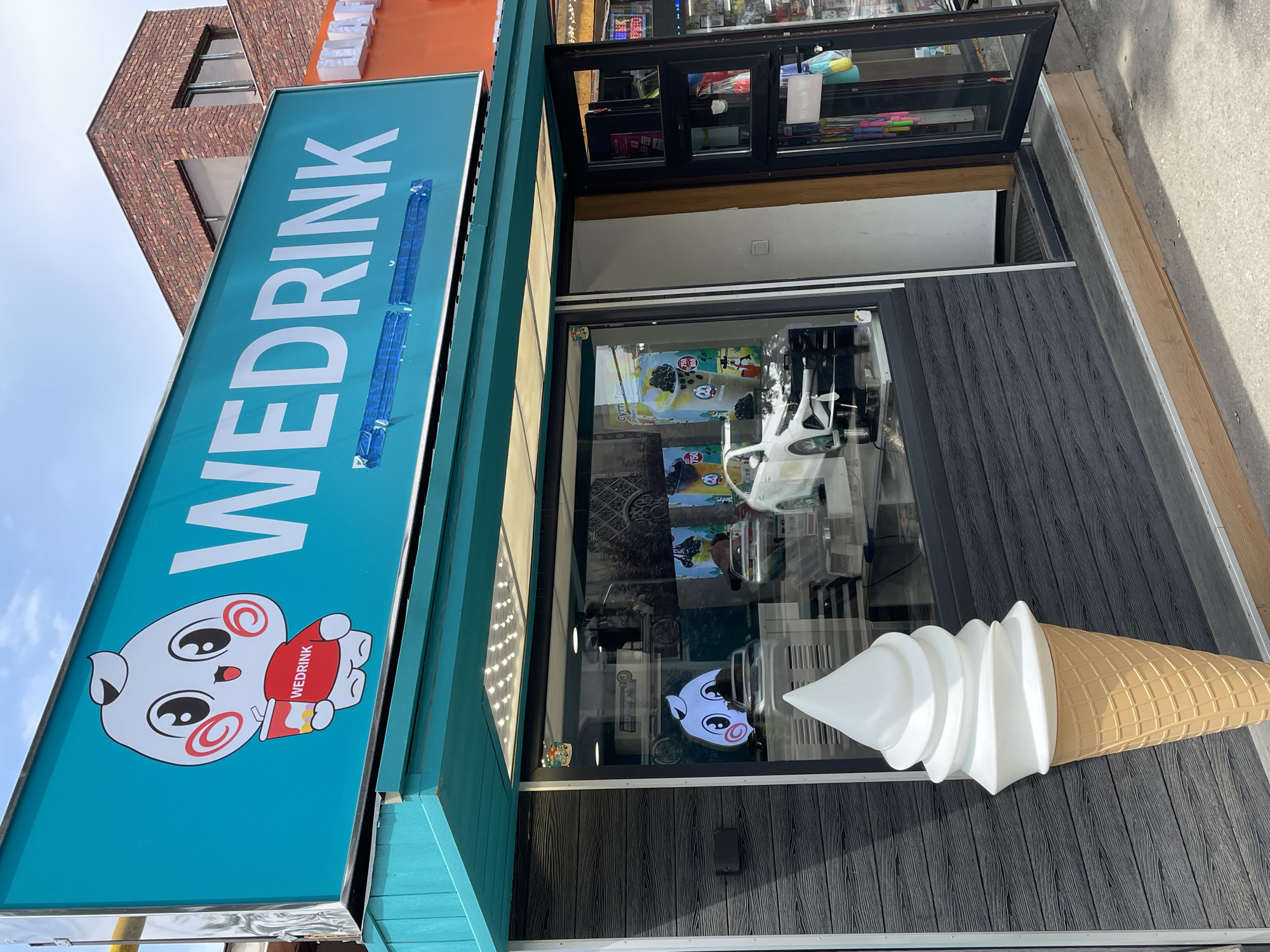 The image shows a storefront in Almaty, Kazakhstan, with a large sign that reads "WEDRINK" in white lettering on a teal background. A cartoon character holding a cup is featured on the left side of the sign. Below the sign, the shop has a large display window showcasing a large, realistic-looking soft-serve ice cream cone sculpture in the foreground. Inside the window, there are reflections and items, including what appears to be a miniature white car and promotional posters. The storefront itself has a dark, textured facade and a teal-colored awning. To the right, a glass door is partially open, revealing the interior of the shop, which appears to sell various items, including what look like jump ropes hanging near the entrance. The overall impression is a modern, brightly colored establishment, likely a cafe or dessert shop, located in an urban environment as indicated by the brick building visible in the background.