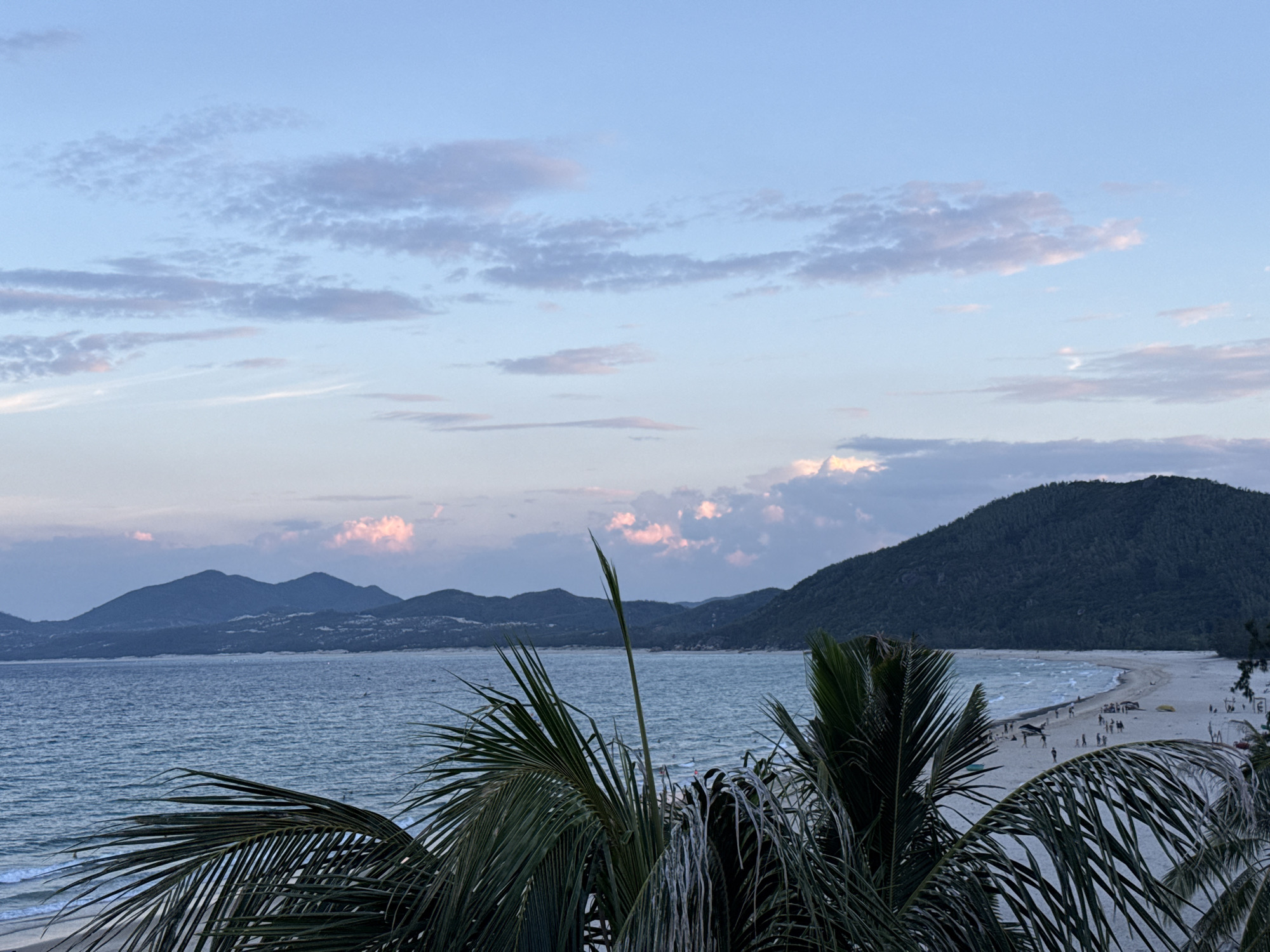 A wide shot captures a tranquil twilight scene on a tropical beach. The sky is a soft gradient of pale blue to lilac, dotted with wispy clouds tinged with pink and orange. A vast expanse of calm, blue ocean stretches across the middle of the frame, with gentle waves lapping at the shore. Along the coastline, a crescent-shaped beach is dotted with small figures of people strolling and interacting. Lush green, mountainous terrain frames the horizon, with one prominent, densely forested hill rising on the right side of the image. In the foreground, the fronds of several palm trees, rendered in dark green, create a naturalistic frame, their leaves partially obscuring the view of the beach and ocean. The lighting suggests either dawn or dusk, casting a serene and peaceful atmosphere over the landscape.