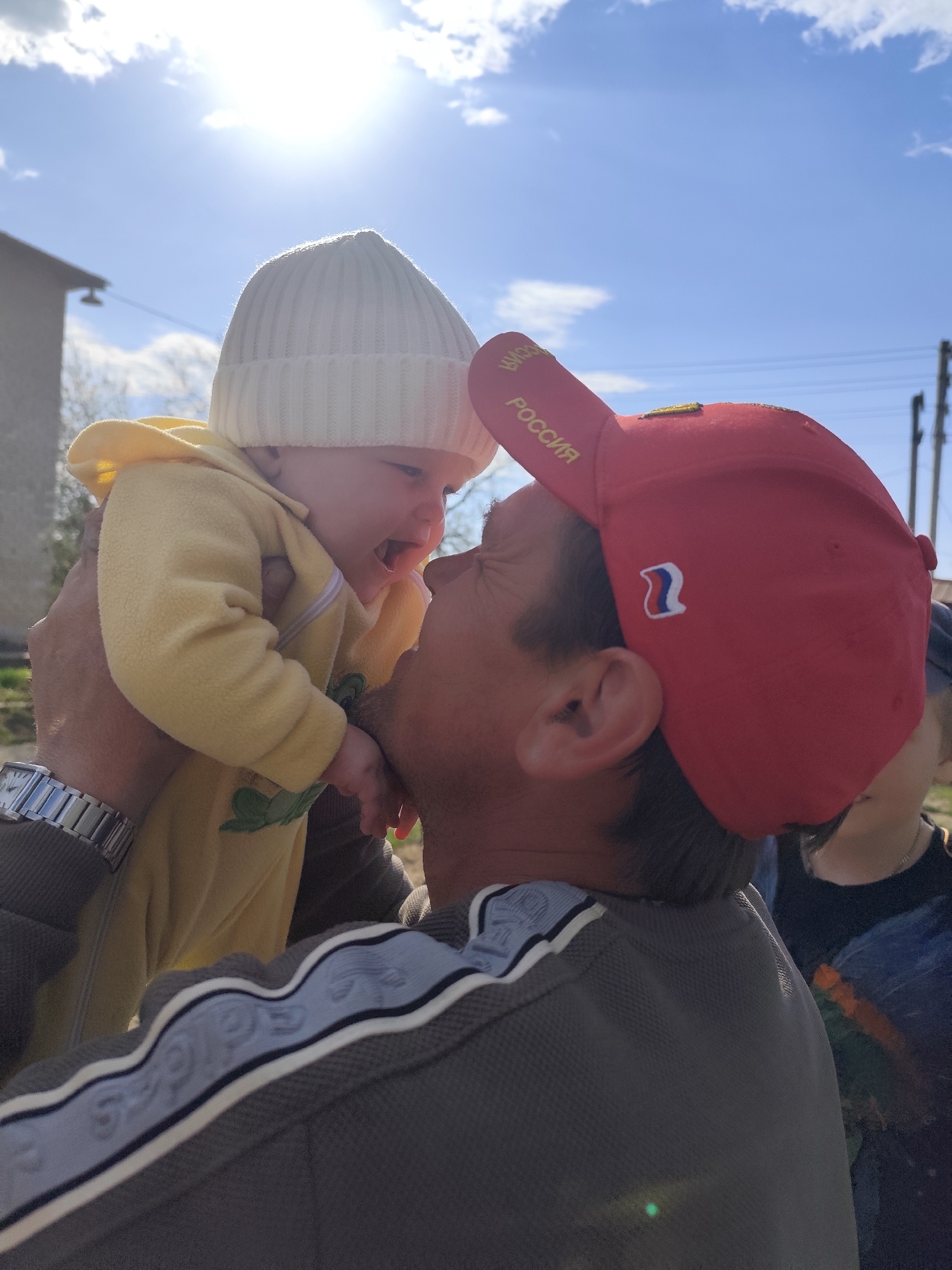 A man wearing a red baseball cap with "РОССИЯ" embroidered on it holds a baby in a yellow outfit and white beanie close to his face. The man wears a grey shirt with blue and white accents. He has a silver watch on his left wrist. The baby appears to be smiling or crying. The background shows a partly cloudy sky, a building, and power lines. A child's face is partially visible in the lower right.