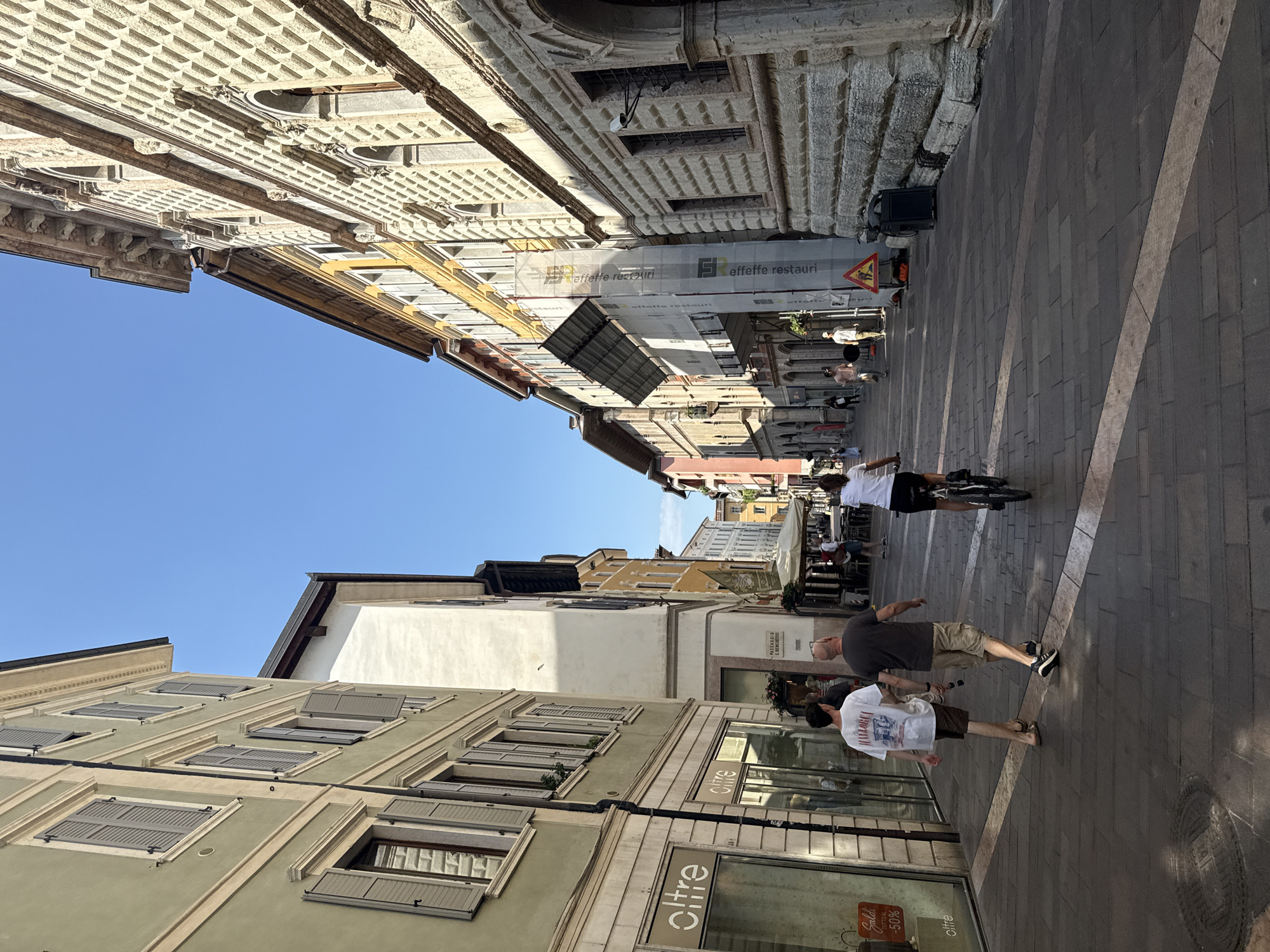 The image depicts a street scene in the city of Trento, Italy, likely on a sunny day given the bright lighting and clear blue sky. The perspective is from street level, looking down a narrow pedestrianized street lined with multi-story buildings. The architecture on the left features buildings with pastel green and white facades, featuring multiple windows with grey shutters. One building has shopfronts at ground level, with the word "oltre" visible on signage and a "Saldi" (sales) sign with "50%" discount displayed.
The right side of the street showcases more elaborate architecture. The closest building has a distinctive facade covered in a diamond-shaped pattern of light-colored stone. Further down, buildings vary in color, with yellow and terracotta hues visible. Some buildings appear to be under renovation, with scaffolding and a large white tarp covering parts of them, bearing text that is partially obscured but includes "effettorestauri".
Several people are present in the scene. In the foreground, two men are walking away from the camera down the street. The person on the left wears a white t-shirt with a graphic print and brown shorts, while the person on the right wears a dark grey t-shirt and khaki shorts. Further down the street, a woman is riding a bicycle, also moving away from the camera. Other individuals are visible walking and standing in the distance, suggesting a moderately busy urban environment.
The street itself is paved with dark rectangular stones, with a lighter colored stripe running diagonally across it. Outdoor seating areas for cafes or restaurants are visible under umbrellas further down the street. The overall impression is that of a charming European city street, likely in its historic center, with a mix of residential and commercial buildings and pedestrian activity.
