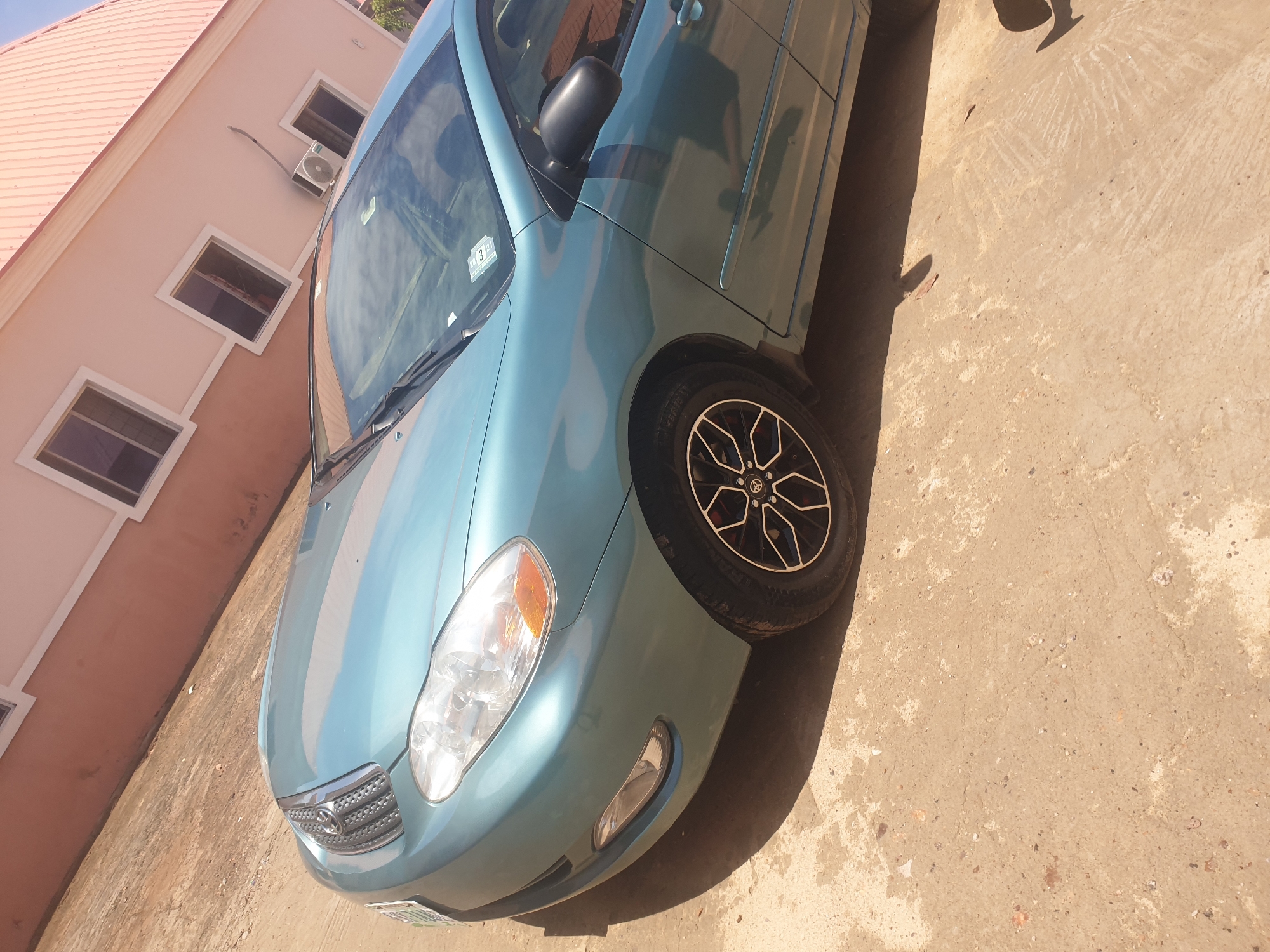 A teal Toyota Corolla is parked on a concrete surface adjacent to a peach-colored building. The building has white-framed windows and an air conditioning unit. The car's front passenger side is facing the viewer. The location is implied to be Lokoja, Nigeria.