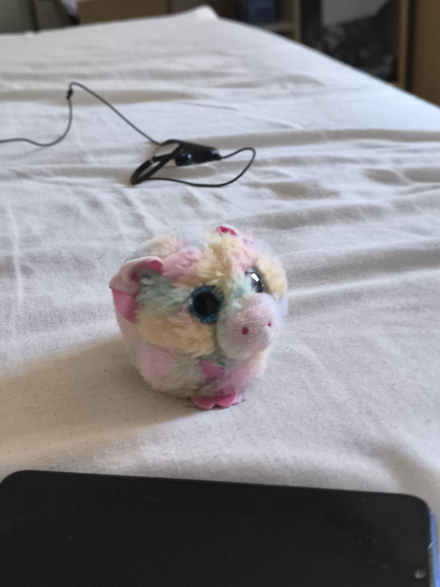 On a white, textured surface resembling a bedspread, a multi-colored plush pig sits in the foreground. The pig features pastel shades of pink, yellow, green, and blue, with large, black, shiny eyes and small, pink ears and feet. A pair of black earphones lies on the bedspread behind the pig. A black smartphone is positioned in the bottom foreground, partially obscuring the lower portion of the image. The background is blurred, showing a wooden shelf and a box. The location in the city of Pélussin, France is unable to be determined.