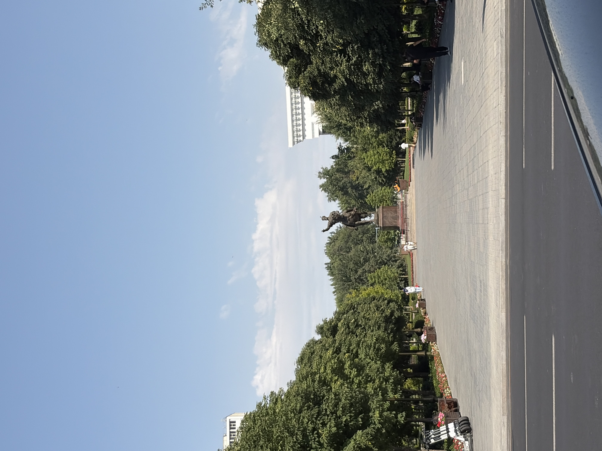 The image shows a wide, paved pedestrian walkway in Tashkent, Uzbekistan, leading towards a large equestrian statue. The walkway is lined with trees on both sides and flower beds. In the background, a white building with ornate detailing is visible on the right, and a modern high-rise building is seen on the left. Several people are walking on the path, and a few are sitting on benches. Electric scooters are parked on the left side of the walkway. The sky is clear blue with some scattered clouds.