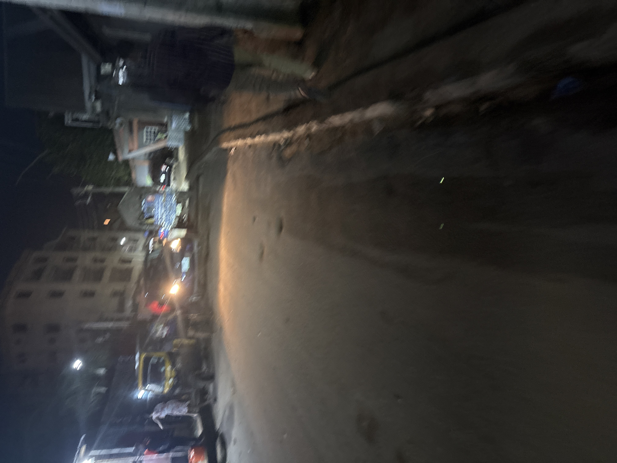 The image depicts a dimly lit street scene at night in Onitsha, Nigeria. A yellow auto-rickshaw is parked on the left side of the road. Several people are visible near the auto-rickshaw, with one person appearing to be interacting with it. Further down the street, a car with its headlights on is approaching. Buildings line the right side of the street, one of which is a multi-story structure. The road surface appears to be asphalt, and there is some debris and vegetation along the right side of the street. The overall impression is of a bustling urban environment after dark.