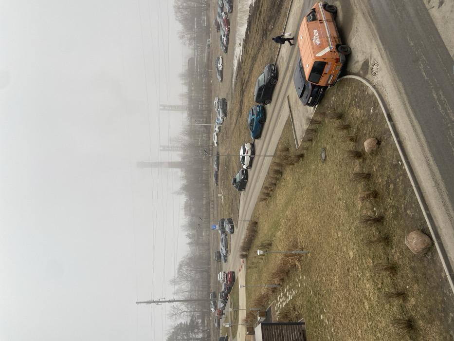 This image depicts a foggy, overcast daytime scene in Новогорелово, Russia, with an industrial background and an urban/suburban foreground.
In the far background, obscured by heavy atmospheric haze, stand multiple tall, brick-colored industrial chimneys, at least three of which are clearly visible, with associated industrial structures. Plumes, possibly steam or smoke, are emitted from at least one chimney. Utility poles and power lines traverse the mid-distance.
The midground features a network of paved asphalt roads and unpaved dirt/muddy areas functioning as parking lots. Numerous vehicles are present; a queue of cars (a black SUV with a roof rack, a white SUV, a blue sedan, and another black SUV) is visible on the paved road in the lower-mid right. An orange commercial van with the word "IEDER" partially visible on its side is parked on the curb in the lower right, behind a black passenger vehicle. Many other cars of various makes, models, and colors (including red, white, grey, and black) are parked both on paved sections and scattered across the unpaved, muddy ground. A small parking sign with a white 'P' on a blue background is visible near the center-left.
The ground cover is predominantly dormant, light brown grass, interspersed with patches of bare earth and mud. Sparse, leafless deciduous trees and low, dormant shrubs line the roads and unpaved areas. A paved pedestrian pathway, bordered by low shrubs and containing at least four streetlights, runs parallel to the main road in the foreground-left. Two large, light-colored boulders are positioned as landscape features in the lower-mid left. Part of a building, featuring a patterned stone patio, is visible in the extreme lower-left corner.
One individual, dressed in dark clothing, walks away from the camera on the right side of the paved road, carrying a white bag. The sky is uniformly light grey, indicating heavy cloud cover and haze, resulting in diffused lighting.