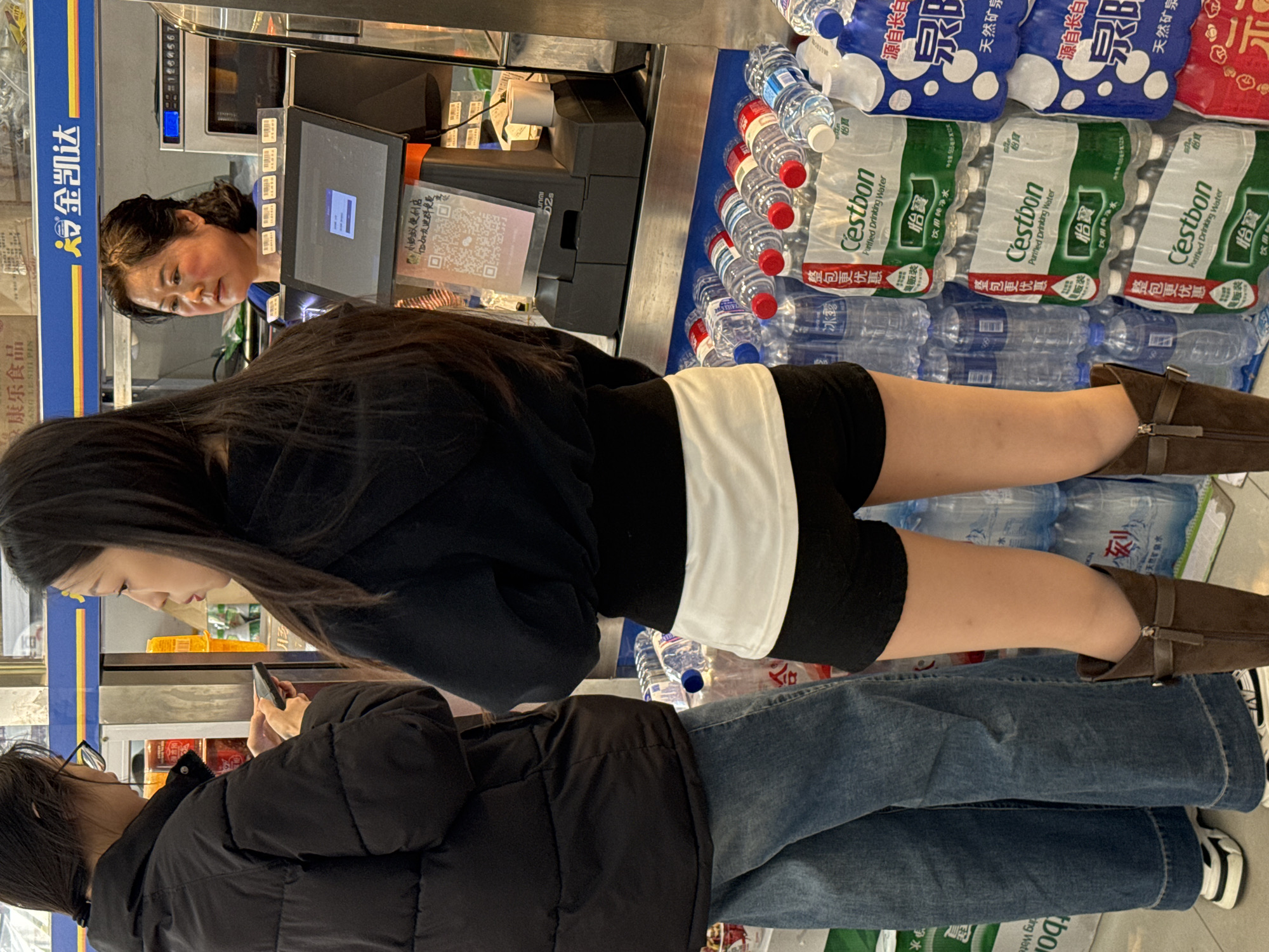 The image shows two young women at the checkout counter of a convenience store, likely in Zhongtun, China. One woman, wearing a black jacket and jeans, is using her phone while waiting. The other woman, dressed in a black top, black shorts with a white stripe, and brown boots, stands closer to the checkout. A cashier, partially visible behind the counter, looks at the customer. The counter is filled with bottled water, including brands like C'estbon and Ice Dew. The store displays Chinese characters and product information.