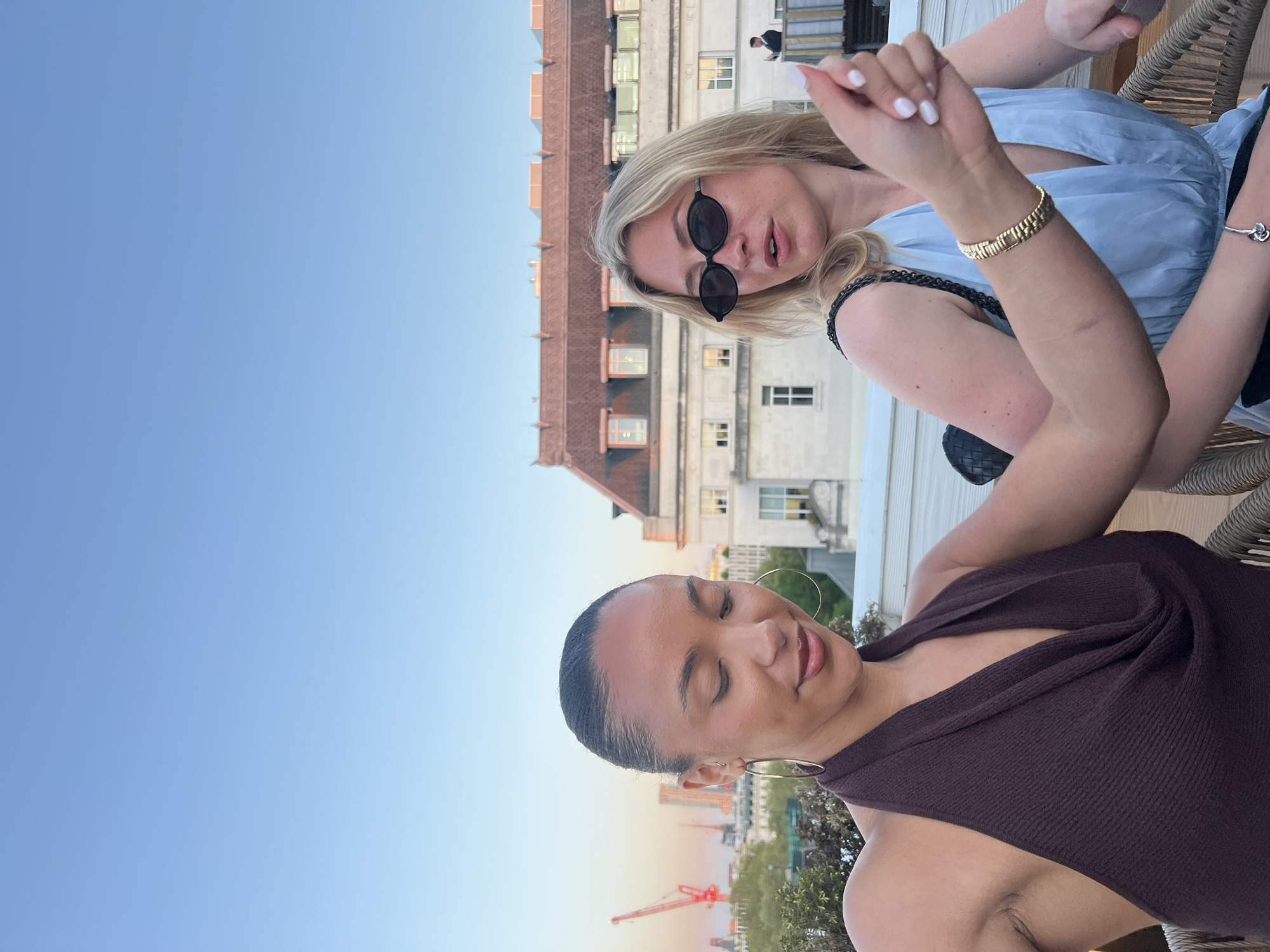Two women are seated at an outdoor venue with a view of London. One woman has fair hair, is wearing black sunglasses and a blue top and a gold bracelet. The other woman has dark hair, large gold hoop earrings, and a brown top. Behind them is a building with windows. There is a crane in the distance. The sky is light blue.