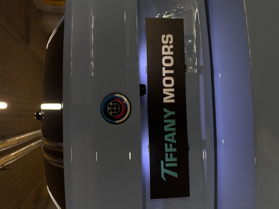 The image displays the rear of a light blue BMW vehicle, likely an M model, parked indoors in what appears to be a parking garage or showroom in Warsaw, Poland. A BMW M heritage logo, featuring concentric semicircles in blue, violet, and red surrounding the standard BMW emblem, is centered on the trunk lid. Directly below this, a black rectangular panel with white lettering reads "TIFFANY MOTORS." The word "TIFFANY" is rendered in a distinct teal font with stylized capital letters, while "MOTORS" is in a standard white sans-serif font. This panel is illuminated from above by a cool-toned white-blue light, consistent with license plate illumination. A small, black rearview camera lens is visible just above the "TIFFANY MOTORS" panel. The car features a dark tinted rear window and a black shark fin antenna on its roof. The indoor environment includes a ceiling with exposed cylindrical ventilation ducts and multiple rectangular, warm-toned overhead lights. The background walls appear to be constructed from a light brown or yellowish-tan material. No individuals are present in the frame.