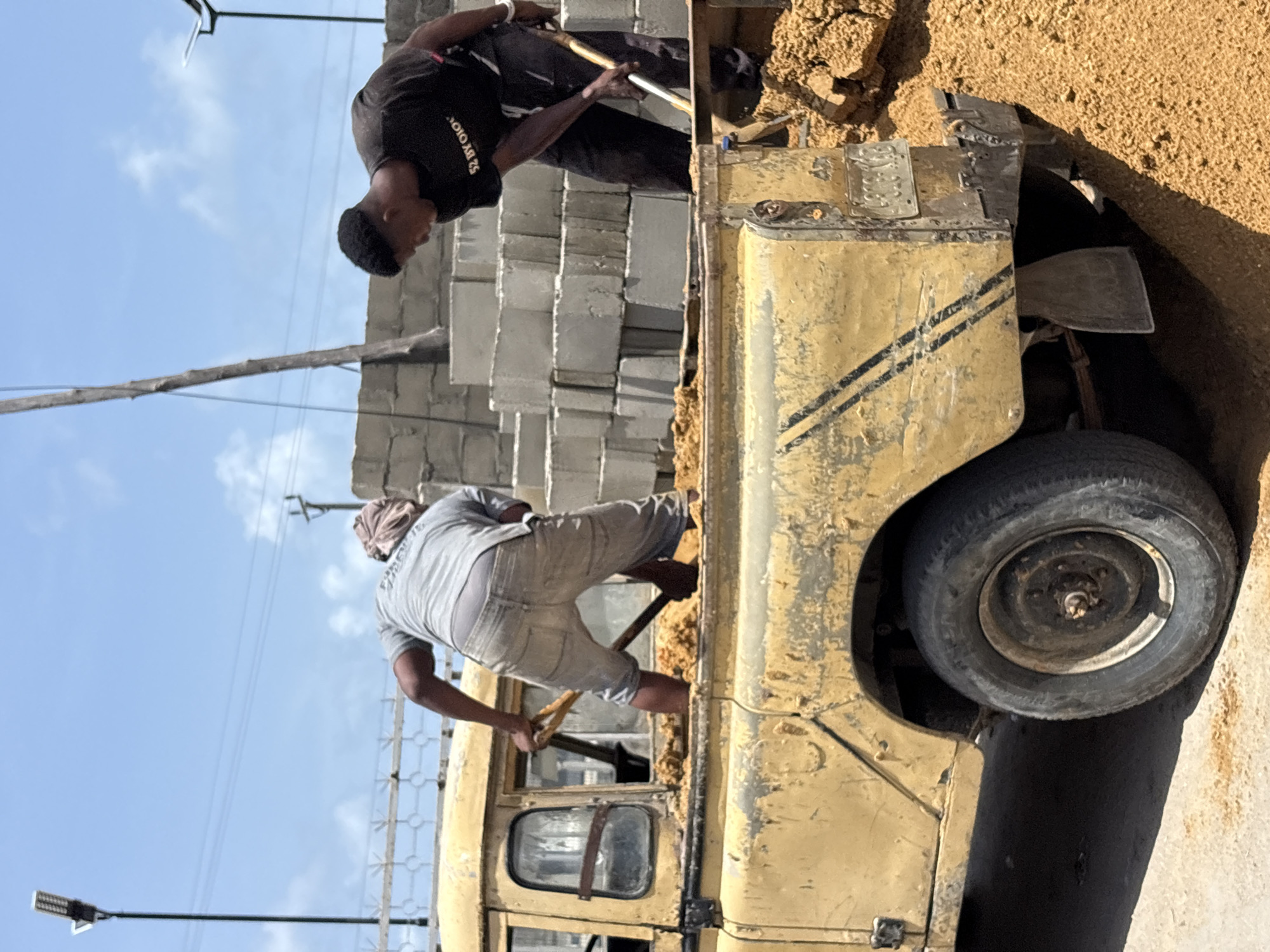This image, taken in Igboefon, Nigeria, depicts two men engaged in manual labor, likely loading or unloading a vehicle with sand or dirt. The scene is set outdoors under a bright, partly cloudy sky during the daytime.
In the foreground, a rugged, yellow utility vehicle, possibly an old pickup truck or jeep, is partially visible. Its paint is weathered with black stripes on the side, and it has a spare tire mounted on the rear. The vehicle appears to be situated next to a pile of sand or light brown aggregate.
Two men are working with shovels. One man, wearing a light grey t-shirt and light-colored shorts, is positioned on top of the vehicle, actively shoveling material. His head is wrapped in a light-colored cloth. The second man, wearing a black t-shirt and dark pants, is standing on the ground near the front of the vehicle, also using a shovel to move material. On his black t-shirt, the text "S2 BY OLOLO" is visible.
In the background, stacks of light grey concrete blocks are neatly arranged, suggesting a construction site or material storage area. A wooden pole and thin wires, possibly power lines, stretch across the sky, and a modern-looking street light fixture on a black pole is also present. The ground is covered in sand and dirt, reinforcing the impression of a construction or transport activity. The overall atmosphere suggests a busy work environment under the open sky.