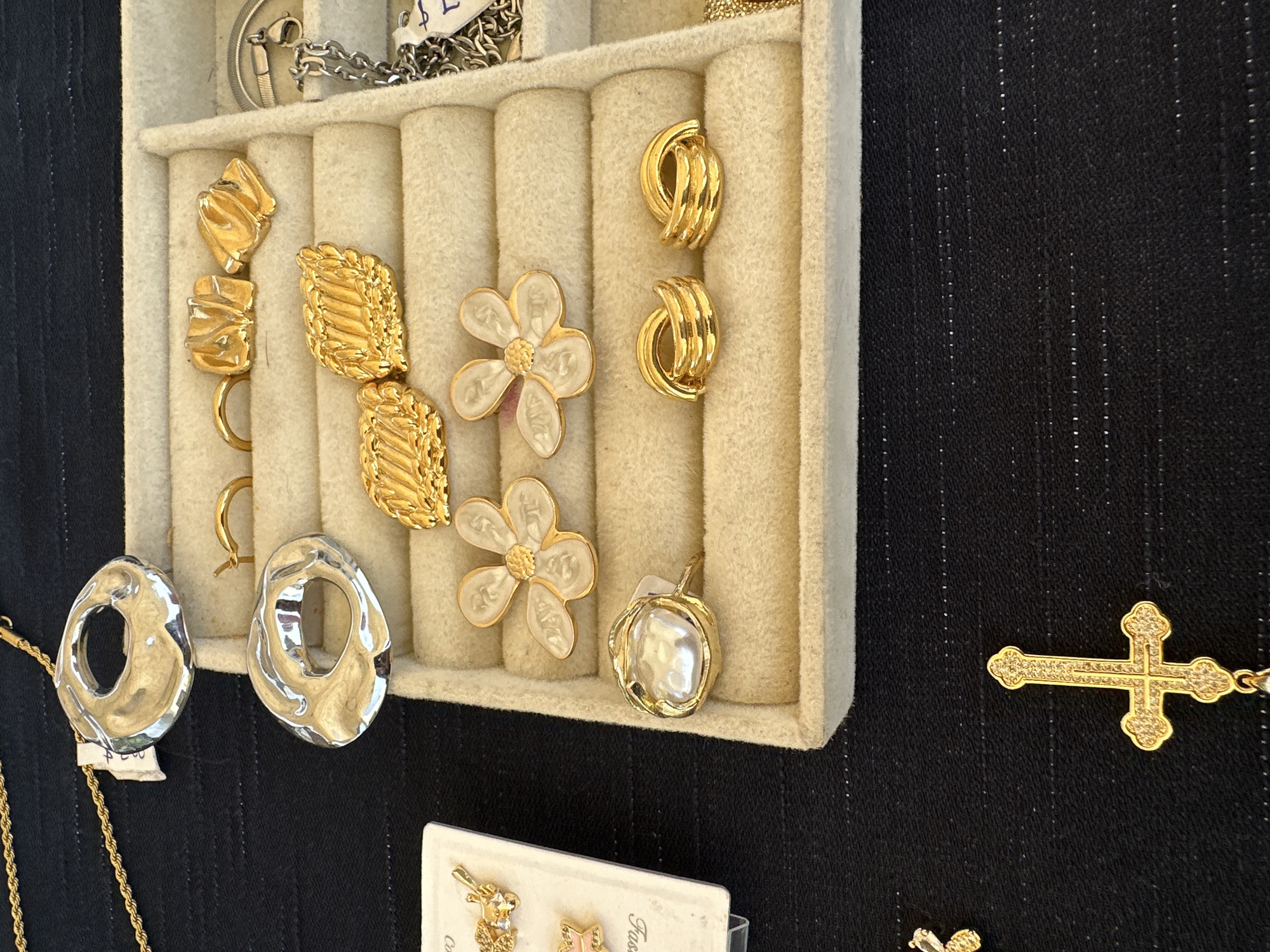 This image shows a display of jewelry, including earrings, necklaces, and a cross pendant, arranged in a velvet-lined tray. The earrings vary in design, featuring gold, silver, and floral patterns. The objects are set against a dark, pinstriped background. Based on the visual context, the scene appears to be a jewelry display or sale within a market or shop. There are no visible people in the frame. There is no reference to location in the image.