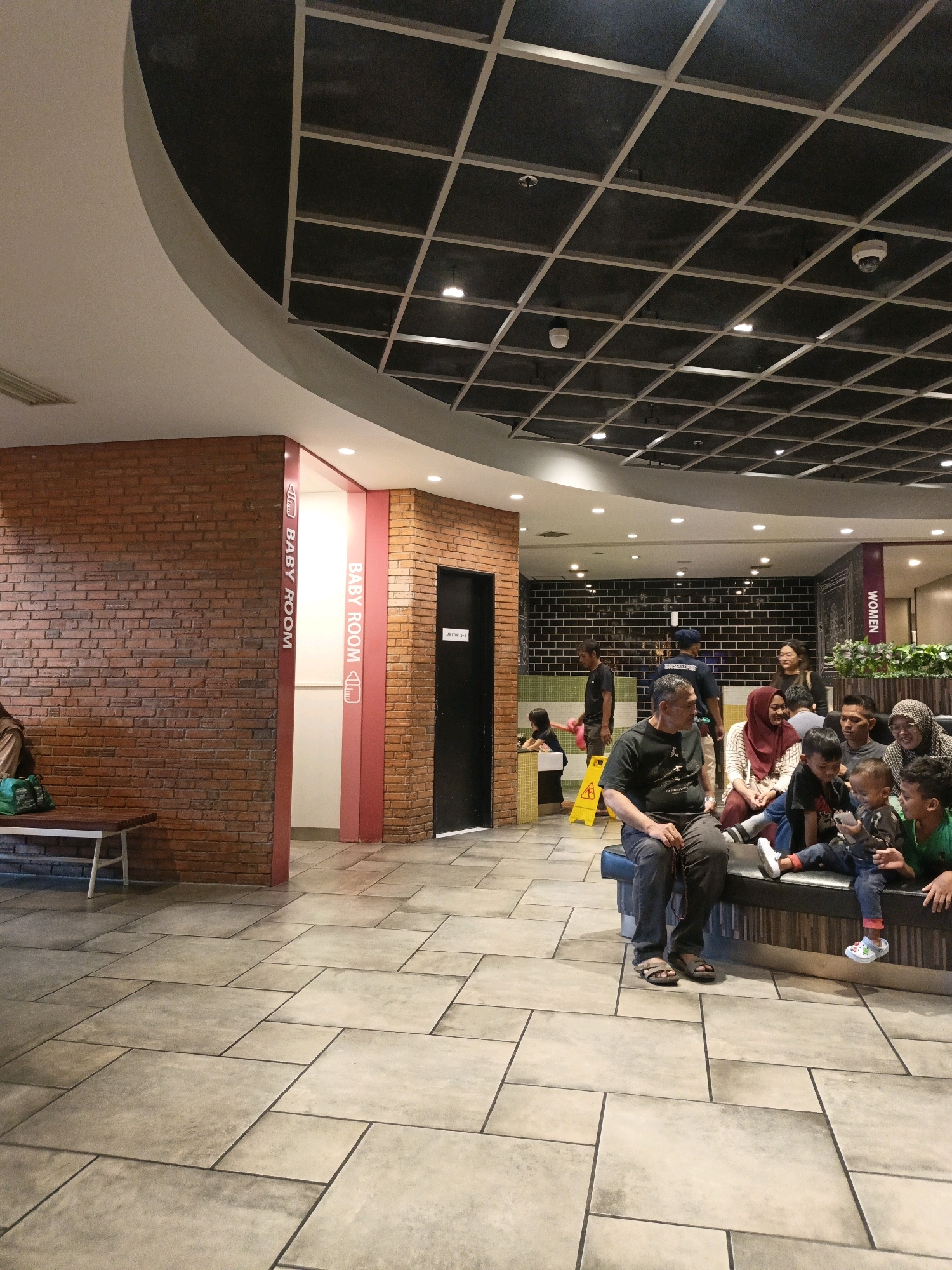 The image depicts an indoor area, likely a public facility such as a mall or restaurant, with a focus on a "Baby Room" entrance and a waiting area. The walls are constructed with exposed red brick on the left and a black tiled wall on the right, adorned with green plants. The floor is tiled with large, rectangular light-colored tiles.
In the foreground, a group of people are seated on a long bench, appearing to be waiting. This group includes several children and adults. A man in a black t-shirt and grey pants is seated on the far left of the bench, facing towards the right. To his right, a woman in a red headscarf is partially visible. Further to the right, a woman in a patterned headscarf is smiling and looking towards the viewer. Several children are positioned in front of the adults, some interacting with each other and one holding a mobile phone.
The "Baby Room" entrance is clearly marked with vertical red panels that have white text reading "BABY ROOM" and a baby bottle icon. Adjacent to this entrance is a black door with white text indicating "UNIT D 3-3."
In the background, a "WOMEN" sign is visible, directing towards another facility. The ceiling features a grid-like structure with dark panels and some lighting fixtures, including what appear to be security cameras. The overall scene suggests a clean and organized public space. The presence of the "Baby Room" and "Women" signs, along with the diverse group of people, indicates this is a family-friendly establishment. There is no explicit indication of the specific location in East Jakarta, Indonesia, beyond the general context of a public building.