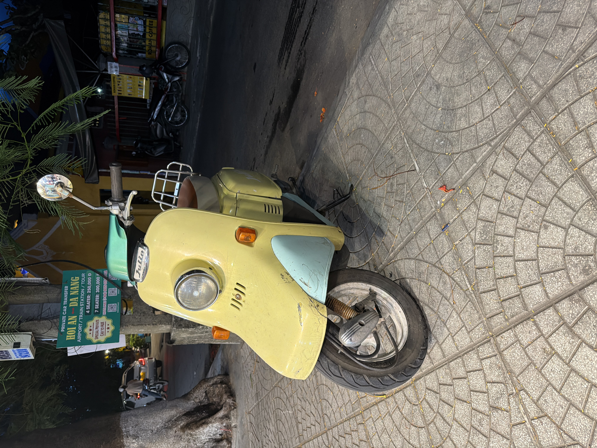 A light yellow and light blue vintage scooter is parked on a sidewalk in Hoi An, Vietnam. The scooter is facing left and its front wheel is visible. The sidewalk is made of gray interlocking pavers with a circular pattern. Behind the scooter, there is a street with a dark surface. In the background, there is a white pole with a green sign advertising "PRIVATE CAR TRANSFER HOI AN - DA NANG AIRPORT/TRAIN STATION/TOUR". There are also buildings and other parked motorcycles visible. A person on a motorcycle is driving away in the background to the left. The scene appears to be at night or in low light conditions.