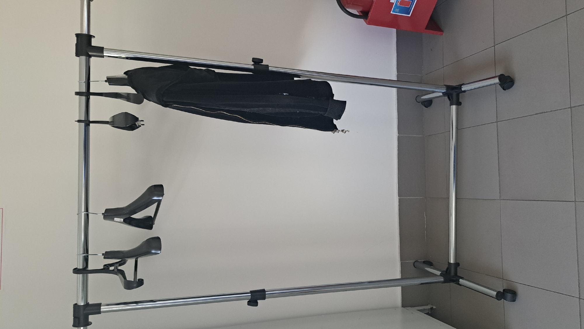 The image shows a portable clothing rack made of chrome-plated metal pipes. The rack is set up in what appears to be a public or commercial space, with a light-colored wall on the left and gray tiled walls on the right. The floor is also tiled.
On the rack, there is a black jacket or coat hanging. Below the jacket, there are three black plastic hangers. Two of the hangers are specifically designed to hold pants or skirts, with clips to secure the garment. The third hanger is a more standard design.
In the upper right corner of the image, a red fire extinguisher is partially visible, mounted on the wall. The lighting suggests it is daytime, and there are no obvious weather cues. The overall impression is a functional space where clothing is being stored or displayed. The setting does not provide specific clues about the exact location within Lytkarino, Russia, beyond it being an indoor environment. There is no visible text in the image.