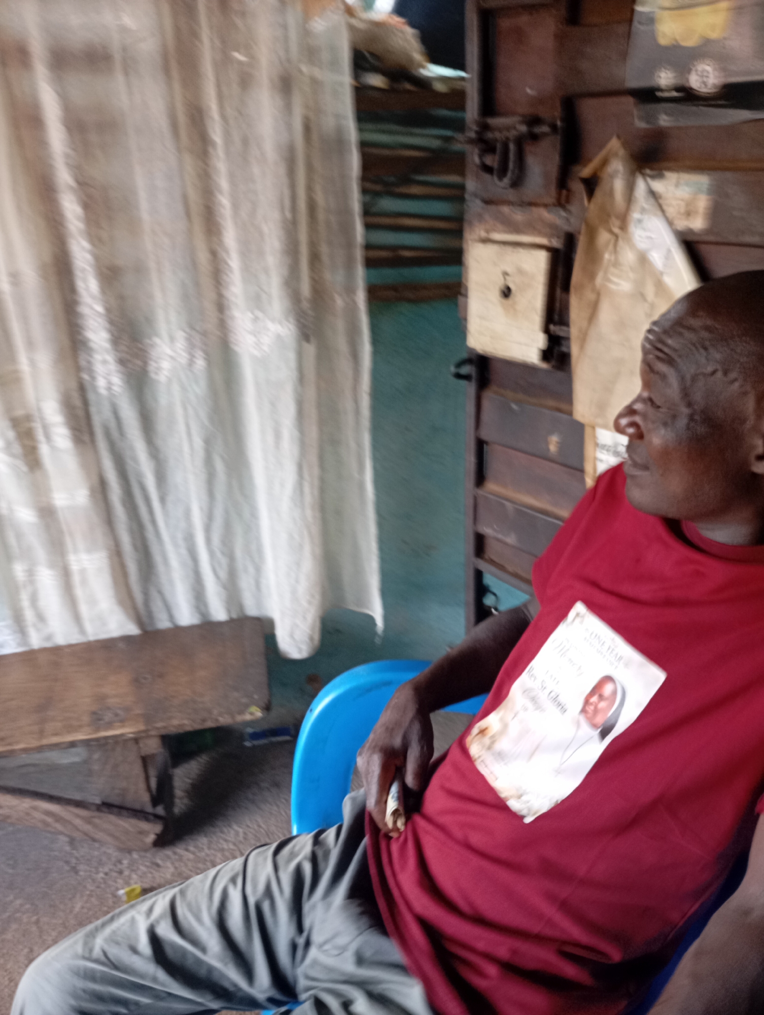 A man wearing a red t-shirt with a memorial image of a woman in religious attire is seated in a blue plastic chair. The t-shirt has text that reads "ONE YEAR ANNIVERSARY" and "Rev. Sr. Gloria". The man is looking to his right. In the background, there is a wooden structure with shelves, a white curtain, and a wooden table. The environment appears to be an indoor setting, possibly a room in a house or a small shop. The image is taken at an angle, and the lighting is somewhat dim.