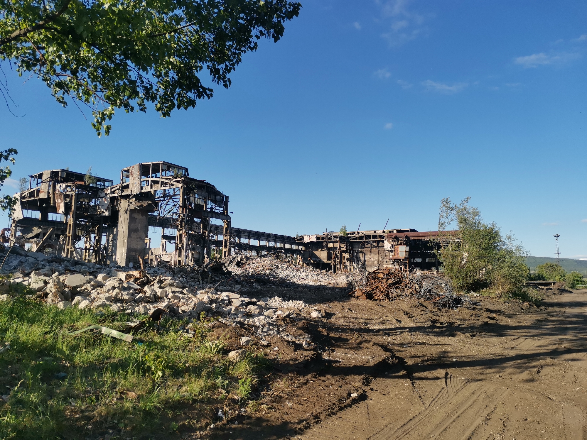 The image depicts the ruins of a destroyed industrial building in Zhivkovo, Bulgaria. The building's skeletal remains are primarily constructed of steel and concrete, with extensive visible damage from fire and collapse. A large pile of rubble and debris lies in front of the building. To the right of the ruins, there is a dirt road with tire tracks, some scattered debris, and vegetation. The sky is clear and blue. In the background, there is a hill or small mountain. A sparse tree is visible in the foreground to the upper left.