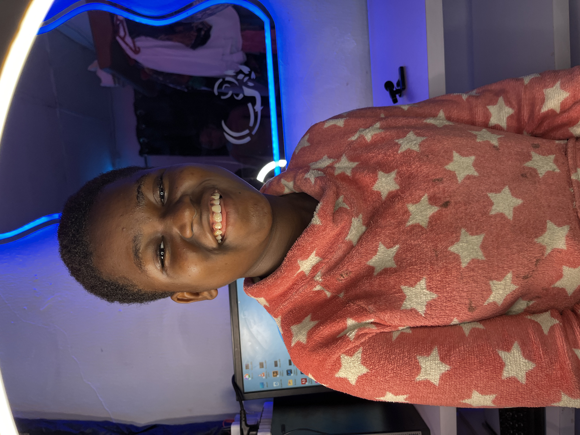 A person with short dark hair and a broad smile is wearing a pink bathrobe patterned with white stars. The person is in front of a computer monitor displaying desktop icons and a curved mirror outlined with blue LED lights. A wall is visible behind the mirror, featuring a black microphone stand mounted on the wall. The location and other details were not discernible from the image.