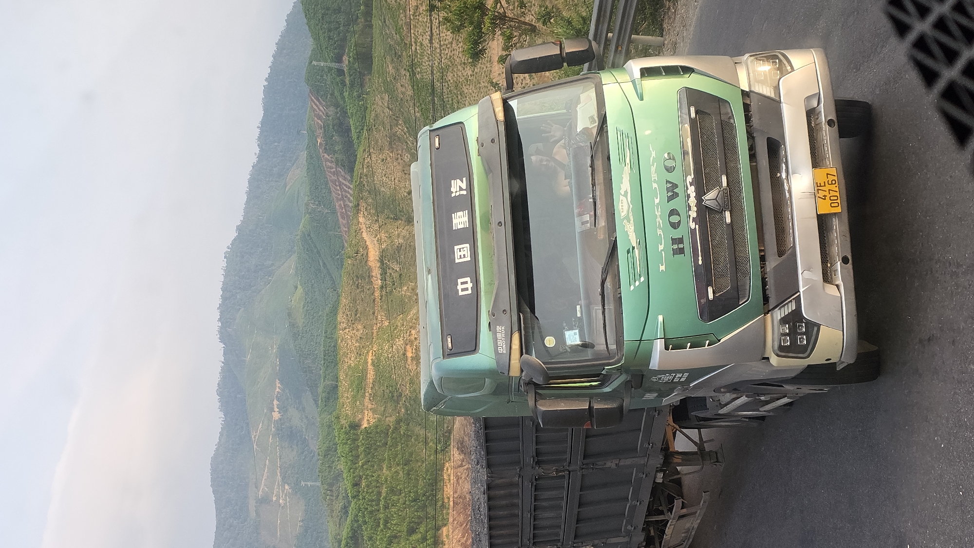 A green and silver HOWO truck is parked on a road. The truck's license plate reads "47E 007.67". In the background, a landscape of hazy green mountains is visible. The scene appears to be located in a rural or mountainous area, with evidence of terraced agriculture on the hillsides. While the image shows a truck, mountains, and road, specific details confirming the location as Aê Thi, Vietnam, are absent.