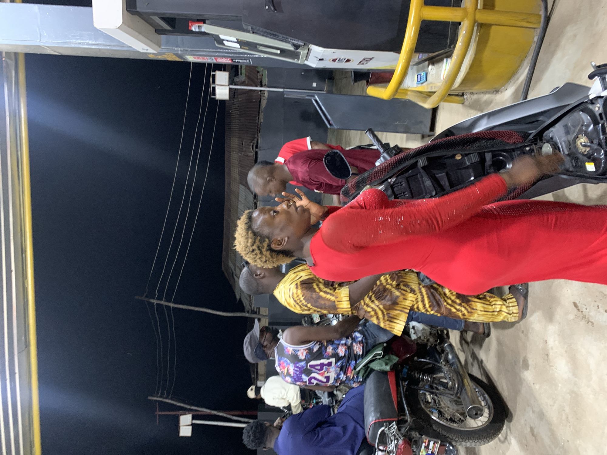 The image depicts a scene at a gas station in Lokoja, Nigeria, at night. Several people are gathered around motorcycles, potentially refueling. One person in a red dress appears to be looking towards another person wearing red. Another person wears a tiger-striped shirt. One motorcycle is near the gas pump, with others parked around it. The background is dark, and the station is illuminated by overhead lights.
