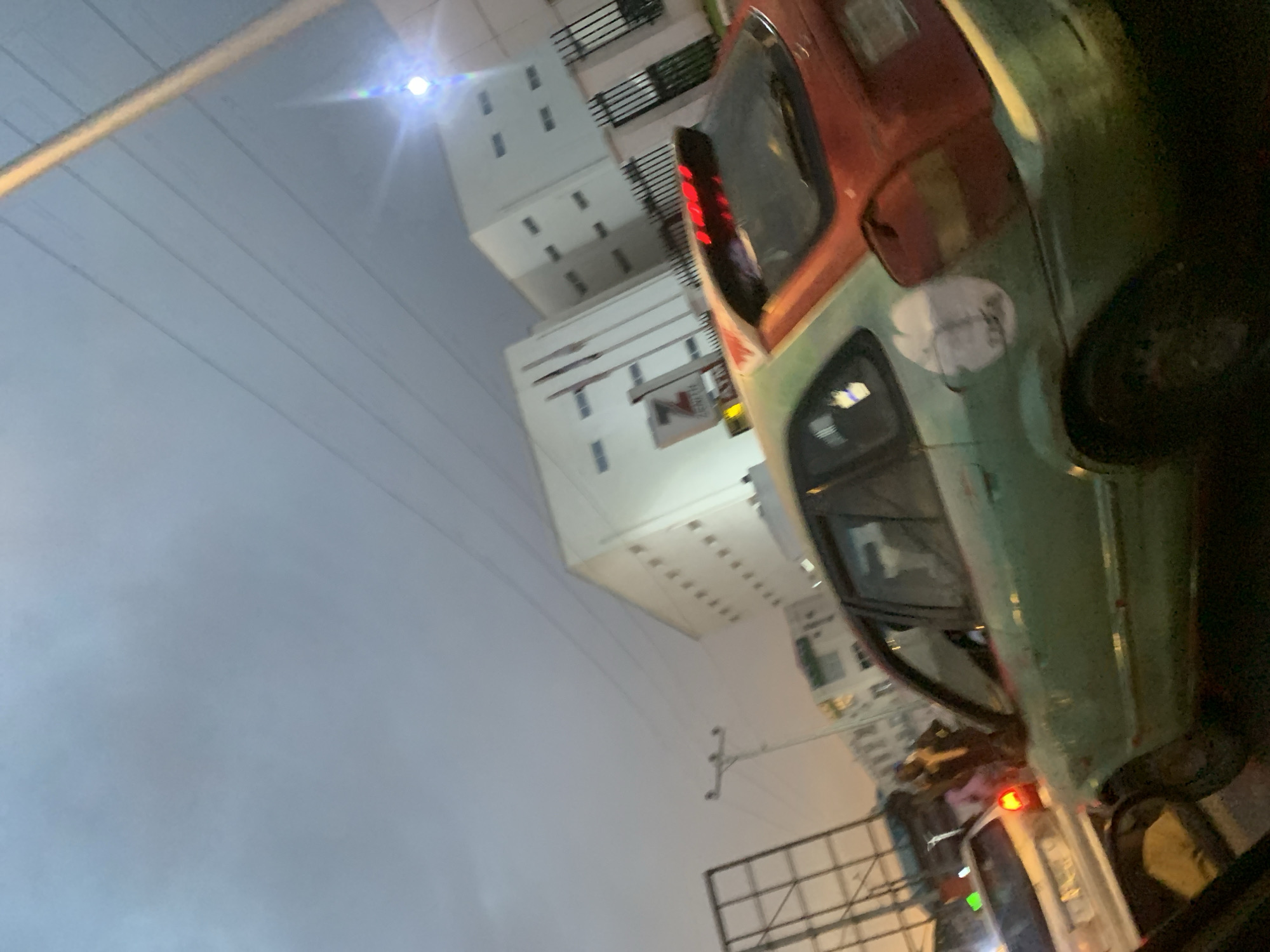 This image captures an urban street scene at night or late dusk. The sky is overcast and hazy, appearing a soft blue-grey.
In the foreground, taking up the right side of the frame, is a large vehicle, possibly a bus or commercial van. It features a distinctive two-tone paint scheme, with a reddish-brown or orange color on its upper half and a lighter green or teal on its lower half. A red taillight is visible at the rear of the vehicle, and a circular white sticker or patch is on its green side. Reflections can be seen in its windows.
To the left and in the midground, several multi-story buildings line what appears to be a street. These buildings are predominantly white or light-colored, featuring numerous small, rectangular windows. On one of these buildings, a sign is visible with the text "ZENITH" alongside a distinct logo, possibly a stylized "Z".
The upper left portion of the image shows a bright light source causing a prominent lens flare. Power lines or utility cables stretch horizontally across the sky. In the lower right, a large metal billboard frame is partially visible, indicating a commercial area. The overall setting is a bustling city environment, typical of a location like Abuja, Nigeria. No people or specific activities are clearly discernible, suggesting a moment of stillness in an urban landscape.