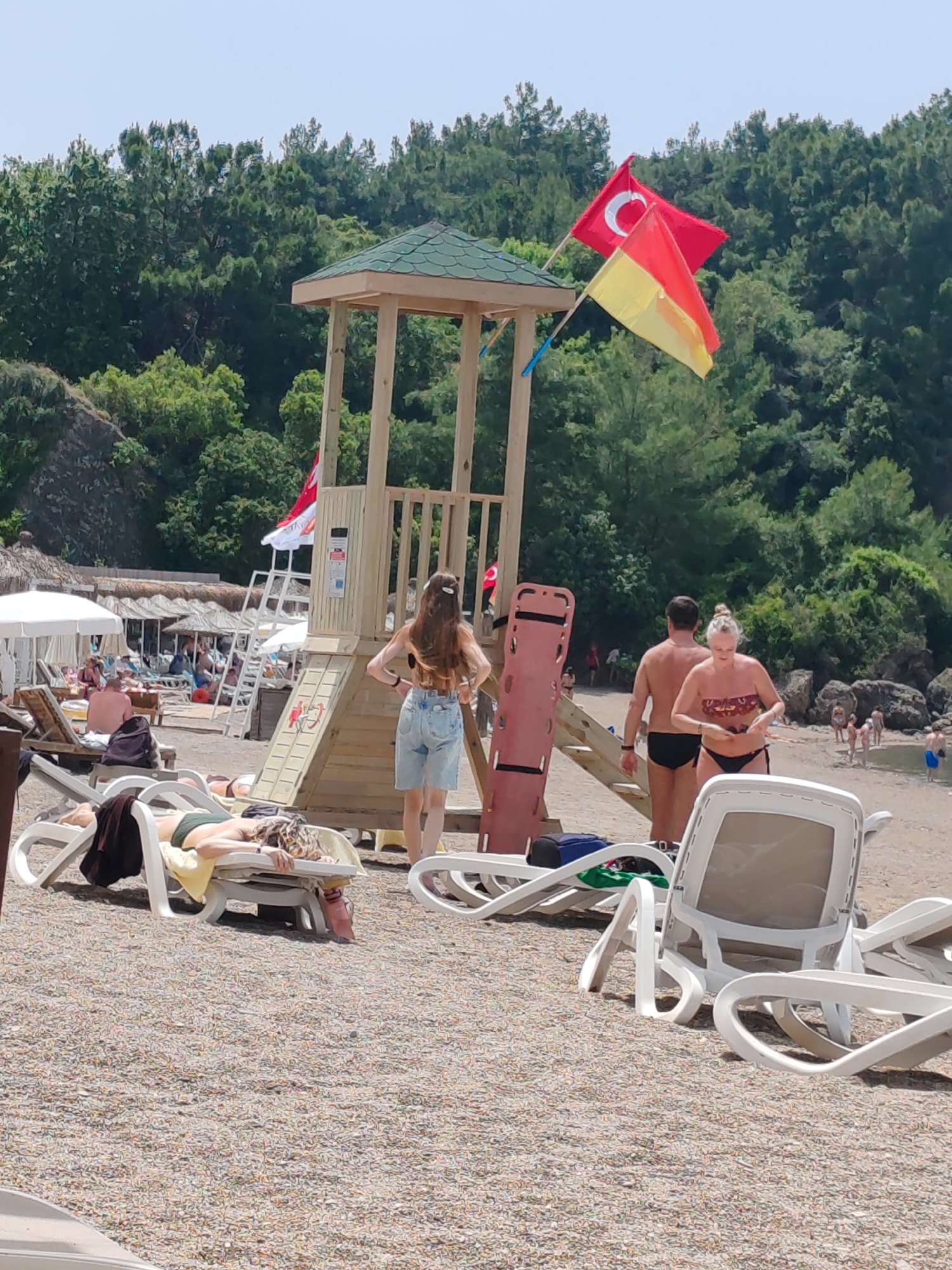 The image shows a beach scene in Alanya, Turkey. A lifeguard tower with a Turkish flag and a yellow and red flag stands on the beach. Several people are present: one woman is standing with her back to the camera, wearing jean shorts and a top; a couple is walking away from the tower, the man shirtless in black swim trunks and the woman in a bikini; another person is lying on a beach chair. In the background, more people are relaxing under white parasols and straw sunshades. White plastic beach chairs are scattered throughout the scene. The beach is covered in pebbles, and a forested area provides a backdrop.