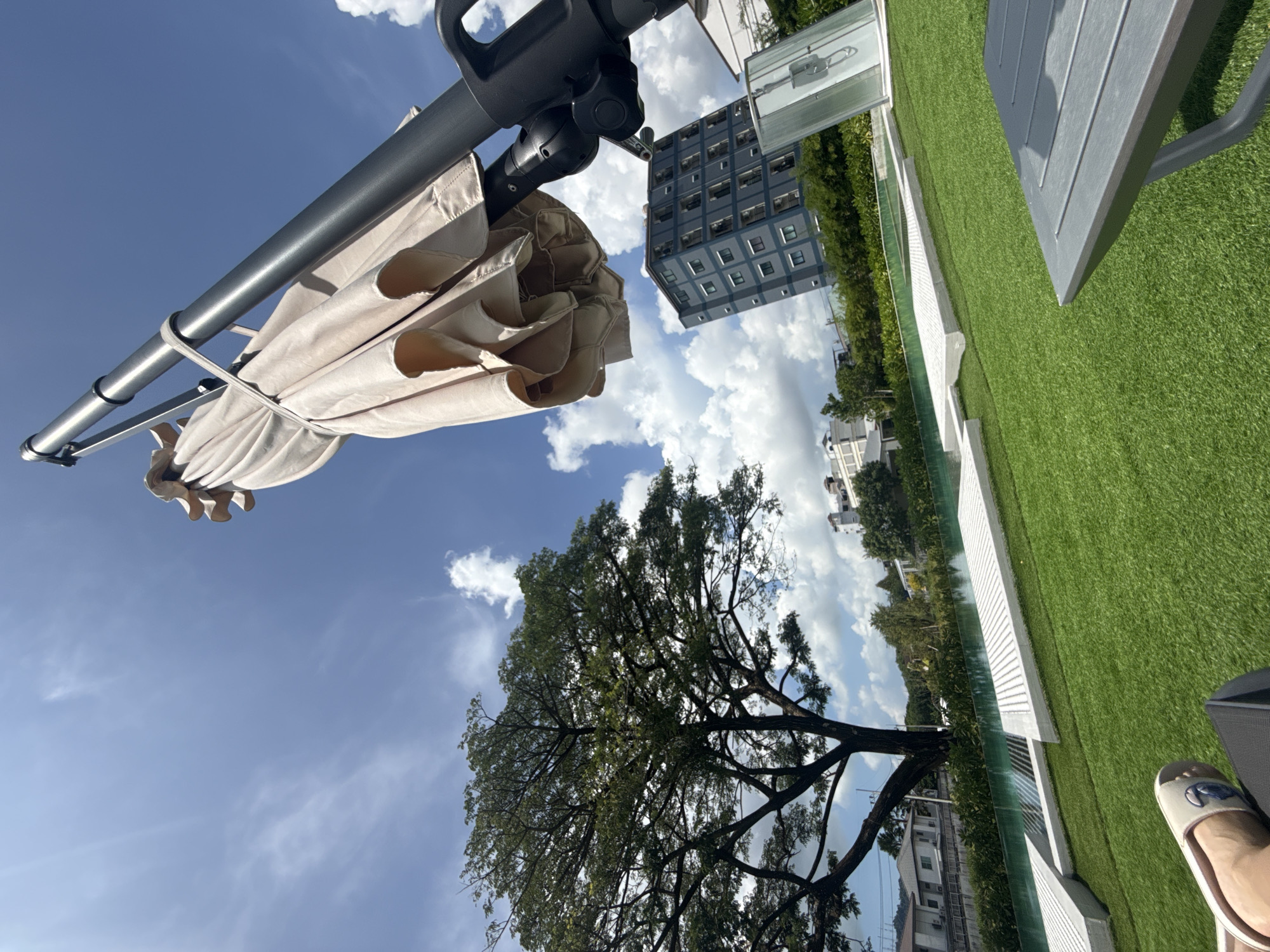 An outdoor scene captures a beige patio umbrella in the foreground, tied shut and angled upwards against a bright blue sky dotted with white clouds. To the left, a large, leafy green tree dominates the middle ground. Behind the tree, a body of water, possibly a swimming pool, is visible, bordered by white structures and lush green landscaping. Further in the background, a modern grey apartment building stands tall, juxtaposed with other residential buildings and more greenery. In the lower right corner, a dark grey lounge chair and a sliver of a person's foot wearing a sandal are visible, suggesting a relaxing moment. The overall setting appears to be a resort or residential complex in the city of Ratsada, Thailand, given the bright sunlight, tropical foliage, and architectural style.