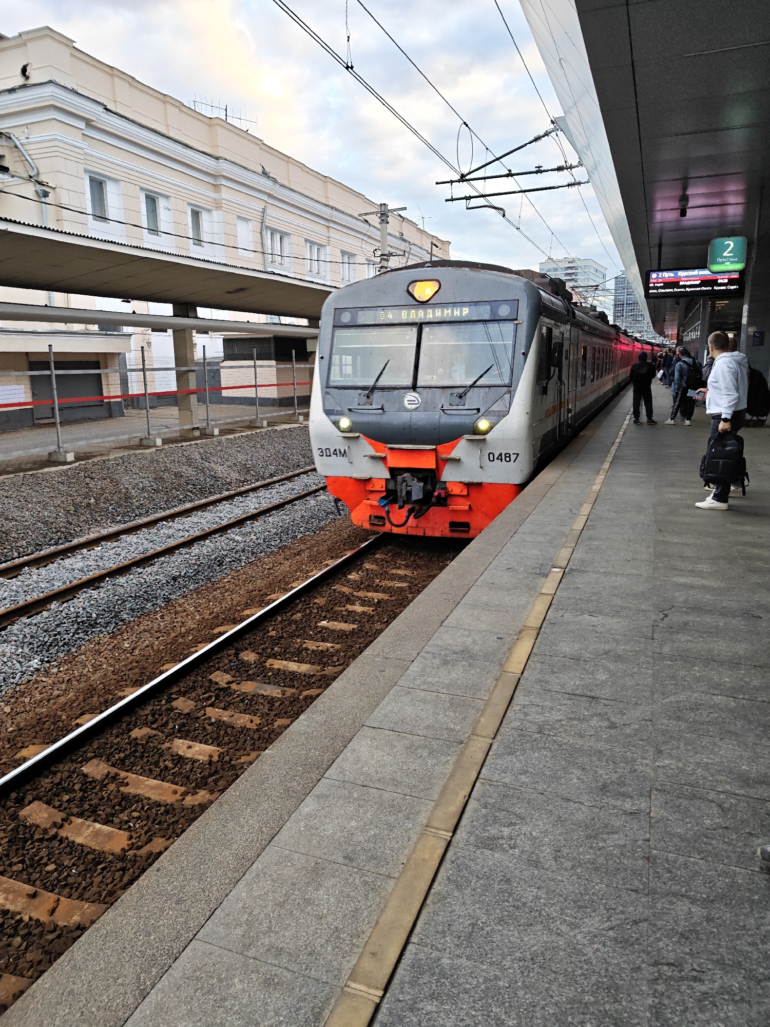 This image shows a train at a Moscow railway station. The train, a 304M electric train numbered 0487, is grey and orange and is stopped at a platform. The station has a modern, covered platform with a digital display showing "2 ПУТЬ" (Platform 2) and train information in Russian. People are standing on the platform, waiting to board or disembark. In the background, an older, light-colored station building with multiple windows is visible. The train tracks are covered with gravel, and overhead power lines for the electric train are strung across the sky. The overall scene depicts a typical urban train station environment.