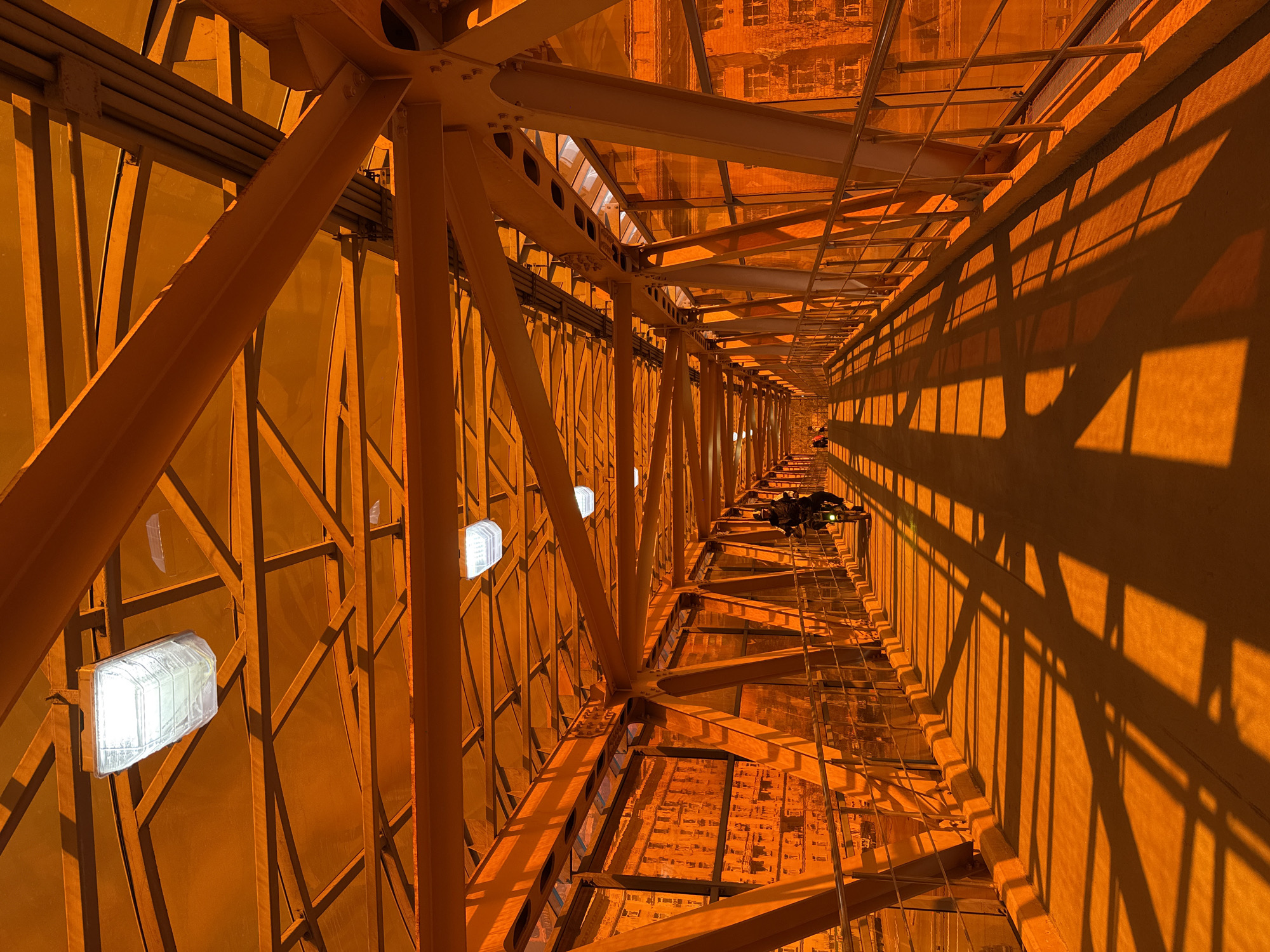 The image depicts the interior of a tower, constructed with orange-painted metal beams and latticework. The structure creates a tunnel-like perspective leading into the distance, with the city of Khimki, Russia, visible at the far end. The internal walls of the tower are illuminated by the setting sun, casting shadows of the internal structure onto the walls. Some light fixtures are attached to the interior structure. In the central distance, two individuals appear to be working atop the tower.