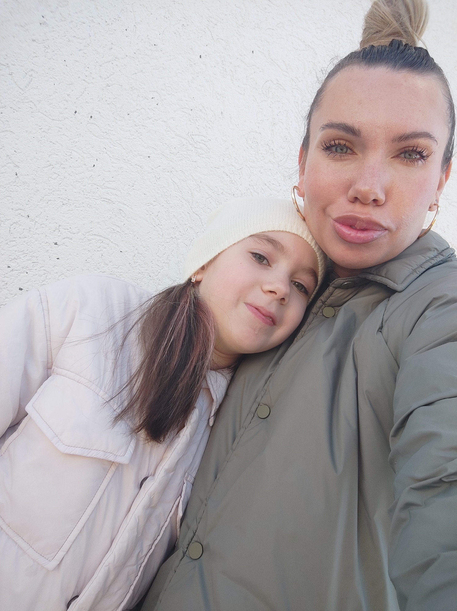 A medium-shot captures a woman and a young girl. The woman, with her hair in a high bun, gold hoop earrings, and a green jacket, is holding the girl close. The girl, wearing a white beanie and a light pink jacket, rests her head against the woman. The background is a plain white wall. The location is not specified but the user requested the city of Votkinsk, Russia as context. No specific objects or actions beyond embracing are visible.