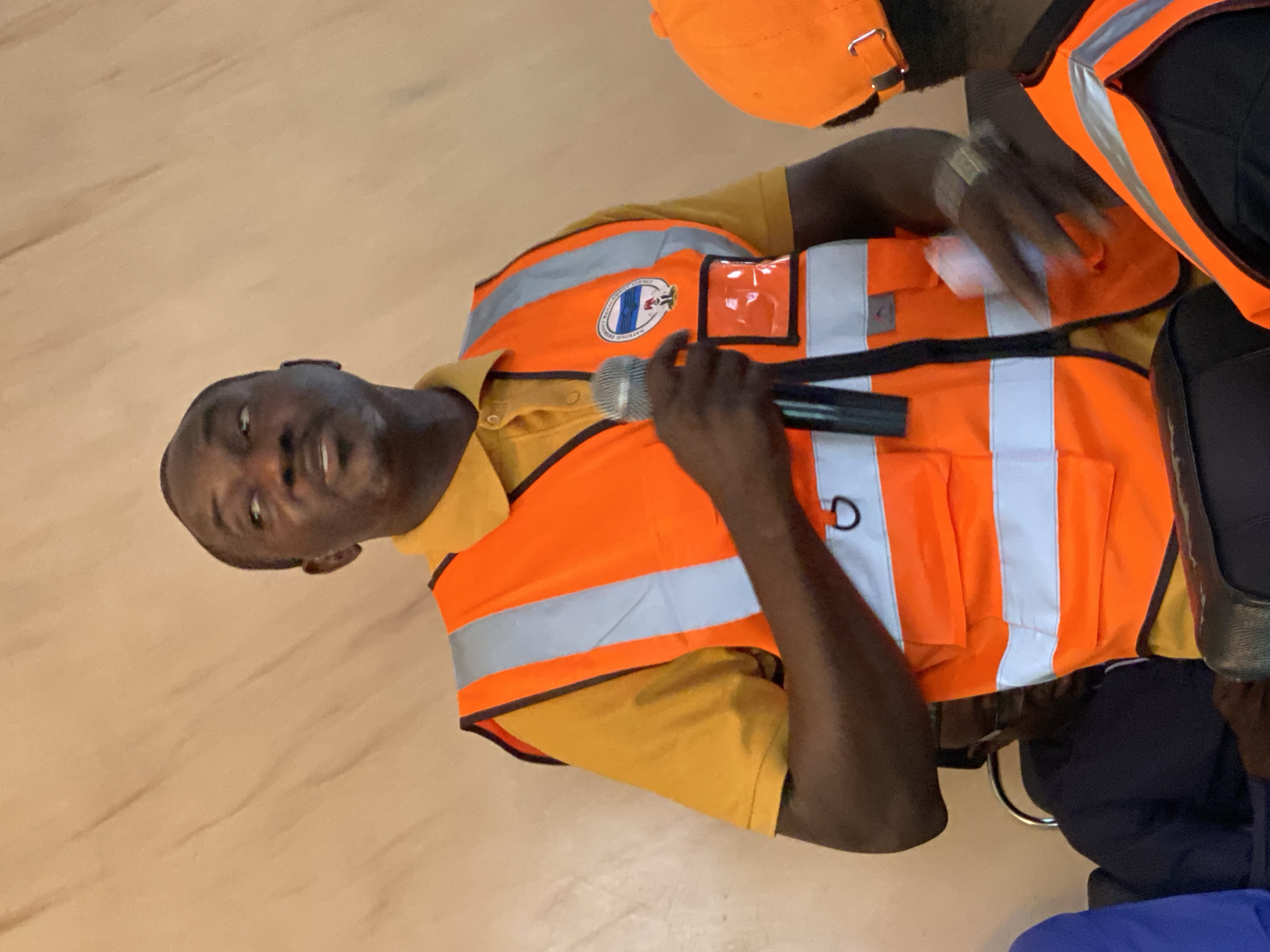 In Unguwar Sarki, Nigeria, a dark-skinned man wearing a bright orange safety vest over a yellow polo shirt speaks into a microphone. The vest has reflective silver stripes and a circular logo on the left chest. He is looking upwards and to his right. His right hand holds the microphone, and his left hand is holding a white object, possibly a piece of paper. Behind him, to his left, another person in a dark shirt and glasses is visible. To his right, a portion of another person wearing an orange cap and a similar safety vest is visible. The background is a textured, light brown wall.