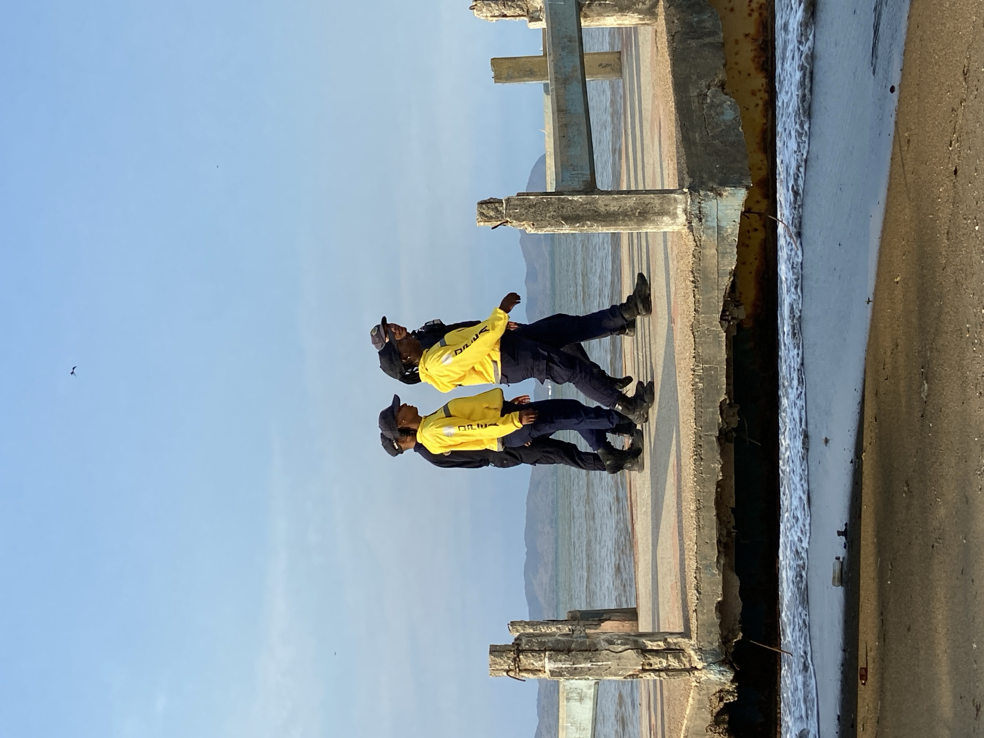 Two individuals in yellow jackets stand on a dilapidated concrete structure overlooking a body of water. They wear dark pants, black shoes, and dark hats. The location appears to be a pier or waterfront structure with weathered concrete pilings and a corroded edge. In the background, there are hazy mountains across the water, and the sky is clear. There appears to be a bird in flight in the distance. This is Higuerote, Venezuela.