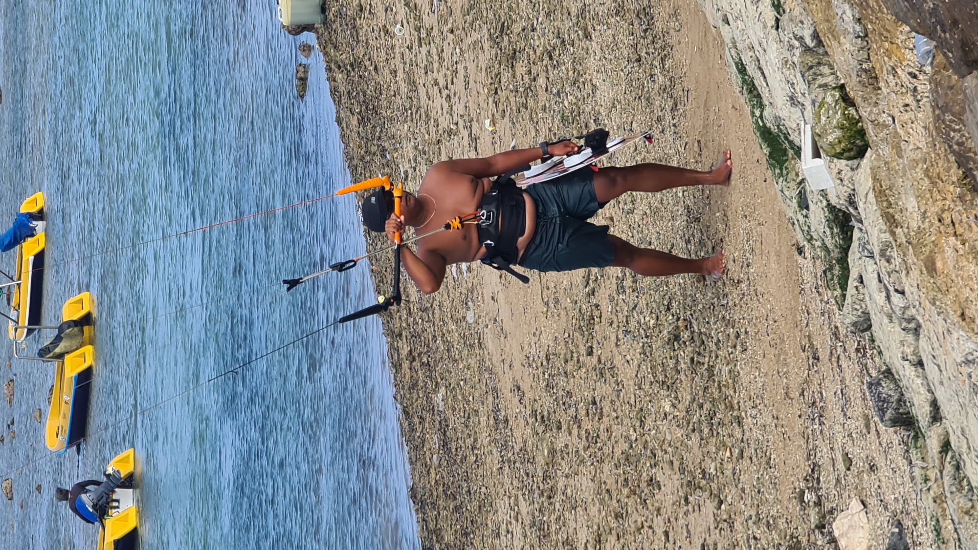 A shirtless man wearing a black baseball cap, sunglasses, and dark green swim trunks stands on a pebble beach, holding a kiteboarding control bar with his right hand and a kiteboard under his left arm. He is wearing a black kiteboarding harness around his waist. The man is facing forward and slightly to his right, looking towards the water. In the background, the ocean is visible, and there are three yellow and blue boats on the water. The beach is covered in small pebbles and some scattered debris. The lower foreground shows a rocky embankment. The scene appears to be a coastal area, likely near Denpasar, Indonesia, given the presence of the boats and the general environment.