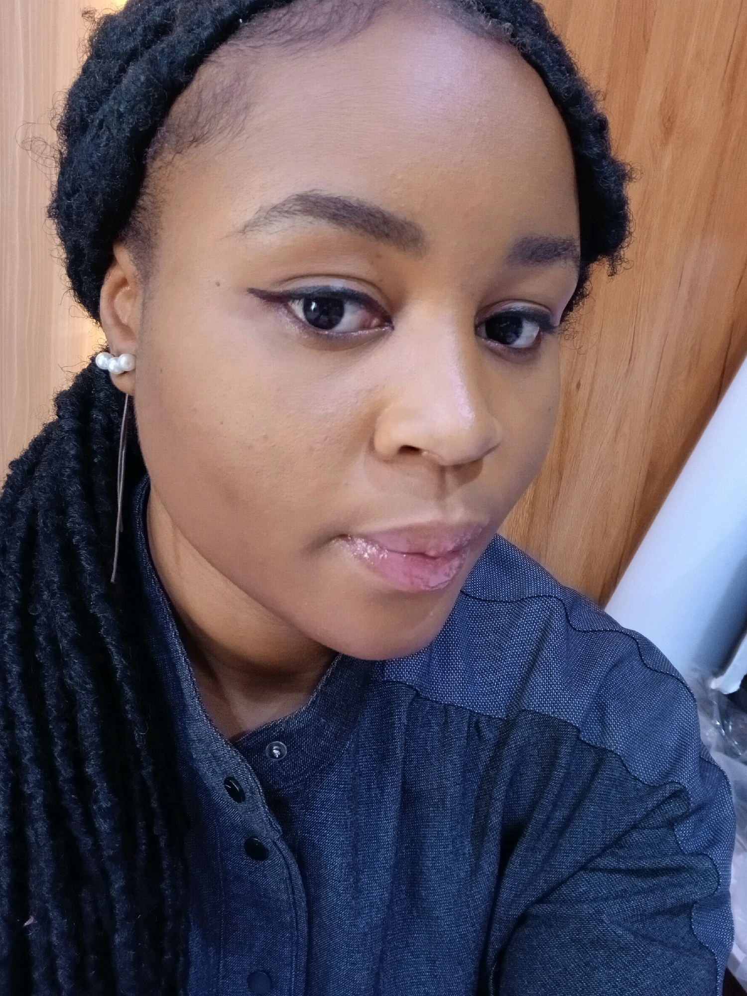 A close-up selfie of a young Black woman is presented, captured from a slightly high angle. The woman has dark skin, dark eyes, and her dark hair is styled in long, thick dreadlocks that fall over her left shoulder. She is wearing a dark blue collared shirt with visible buttons. A delicate pearl earring dangles from her left ear. Her makeup includes dark eyeliner and lip gloss. The background appears to be a wooden surface, possibly a wall or partition, with a sliver of white on the right side of the frame. The lighting is warm and diffuse, casting soft shadows. The image quality is moderate. The provided location of Gura Top, Nigeria is not visually verifiable from the image itself.