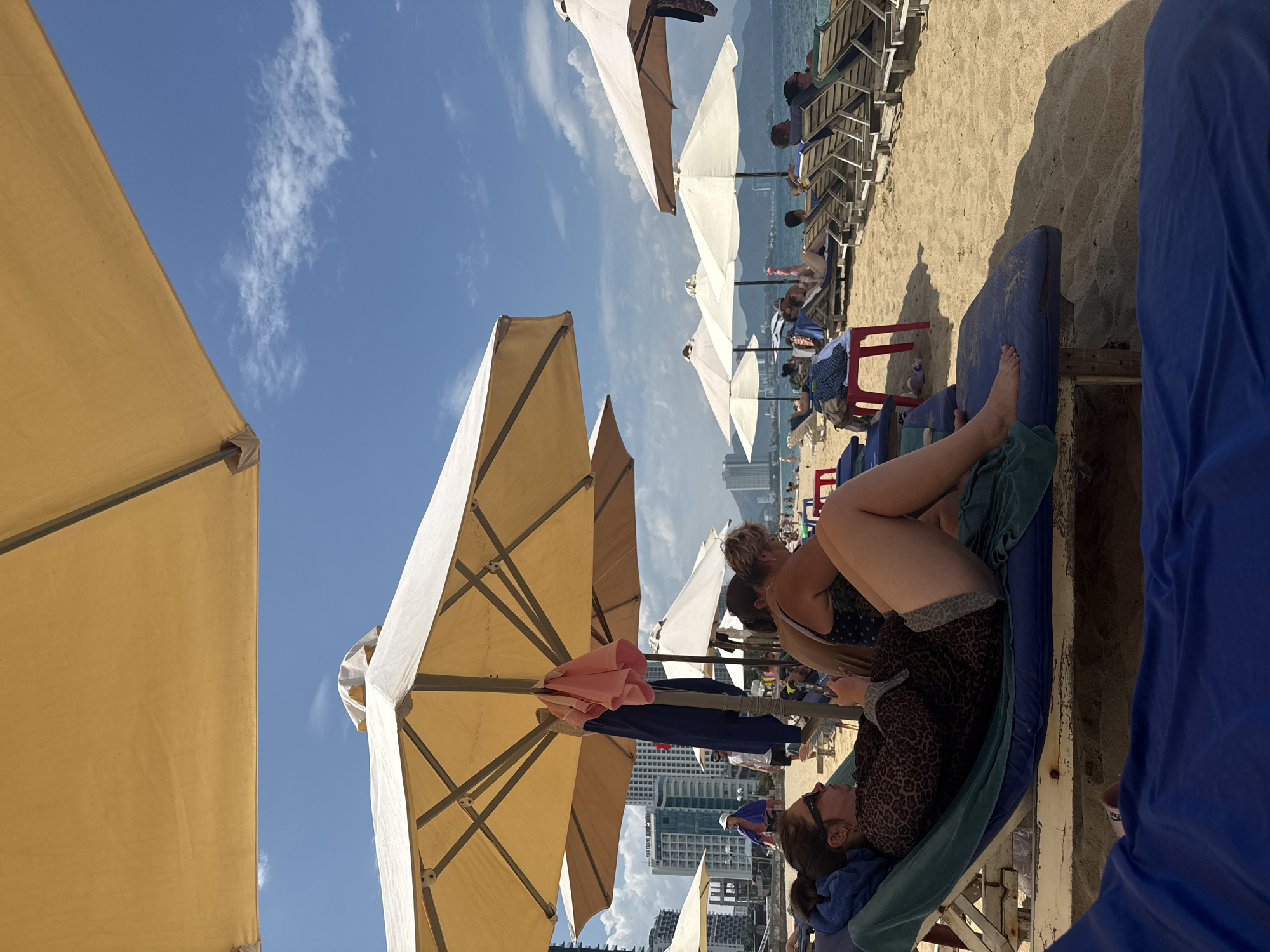 The image captures a sunny day at a beach in Nha Trang, Vietnam. In the foreground, a woman lies on a blue lounge chair, wearing sunglasses and a leopard-print top, holding a phone. The sand is visible, with other beach chairs and umbrellas dotting the scene. In the middle ground, several people relax on lounge chairs under beige umbrellas. In the background, tall buildings and hotels line the city's coastline, with mountains visible in the distance. The sky is blue with scattered white clouds.