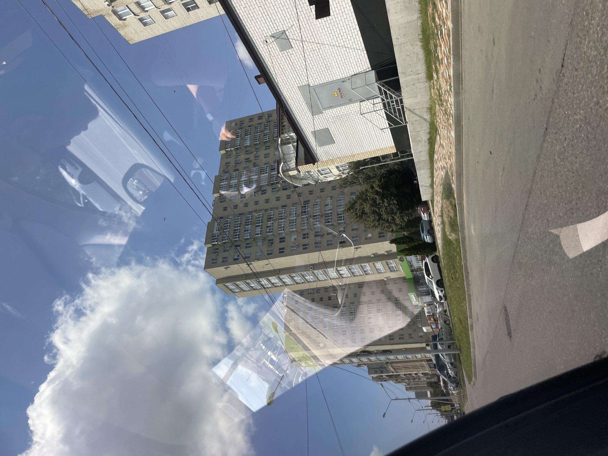The image depicts a street view in the city of Stavropol, Russia, likely captured from inside a moving vehicle due to reflections in the foreground. On the right side of the image, a single-story white brick building with a dark-colored roof and trim is visible. This building has a metal door with a warning sign and a set of stairs leading up to it. To the left of this building, a densely populated residential area is present, characterized by tall apartment buildings with numerous windows. The street in the foreground is paved, with parked cars and grassy areas lining the roadside. Overhead, power lines crisscross the sky, and the sky itself is blue with scattered white clouds. The overall scene conveys an urban environment during daylight hours.