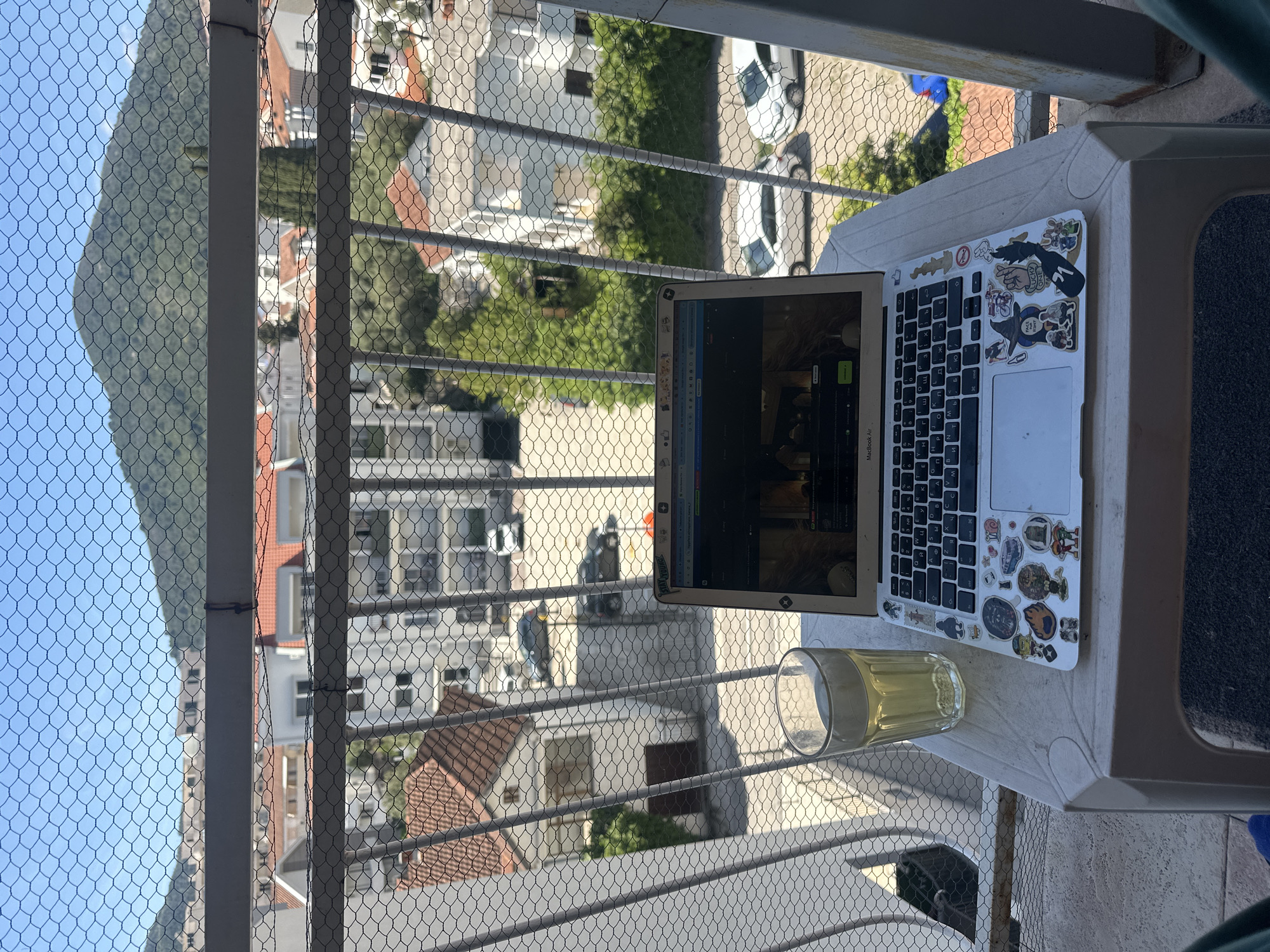 A view from a balcony in Budva, Montenegro, shows a laptop and a glass of yellow liquid on a small white table, with buildings and a mountain visible through a wire mesh railing. The laptop is open, displaying a webpage. Stickers adorn the laptop's lid. The balcony overlooks a residential area with a mix of white and red-roofed buildings, with parked cars visible in the background. A mountain dominates the left side of the frame, beneath a clear blue sky. The perspective is angled downward.
