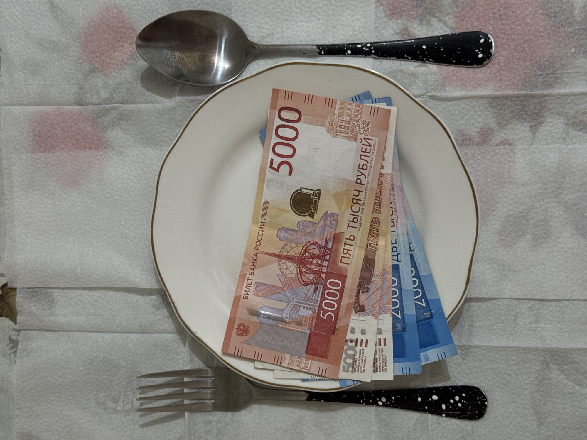 This scene, located in Chaikovsky, Russia, presents an overhead view of a dining table setting. A textured white disposable tablecloth, showing creases, covers a surface that subtly reveals faint reddish-pink patterns beneath. Centered on the tablecloth is a white ceramic plate with a thin gold rim. To the left of the plate, a silver-colored fork with a black handle speckled with white dots is positioned. To the right, a matching silver-colored spoon with a black, white-speckled handle is placed. Both cutlery handles display minor wear and chipped paint. A stack of Russian Ruble banknotes rests on the plate. The uppermost notes are orange-red 5000 Ruble denominations, clearly showing "БИЛЕТ БАНКА РОССИИ," "5000," and "ПЯТЬ ТЫСЯЧ РУБЛЕЙ," along with imagery of Moscow cityscapes. Partially visible beneath these are blue 2000 Ruble banknotes. The entire setup is uniformly lit, casting soft shadows.