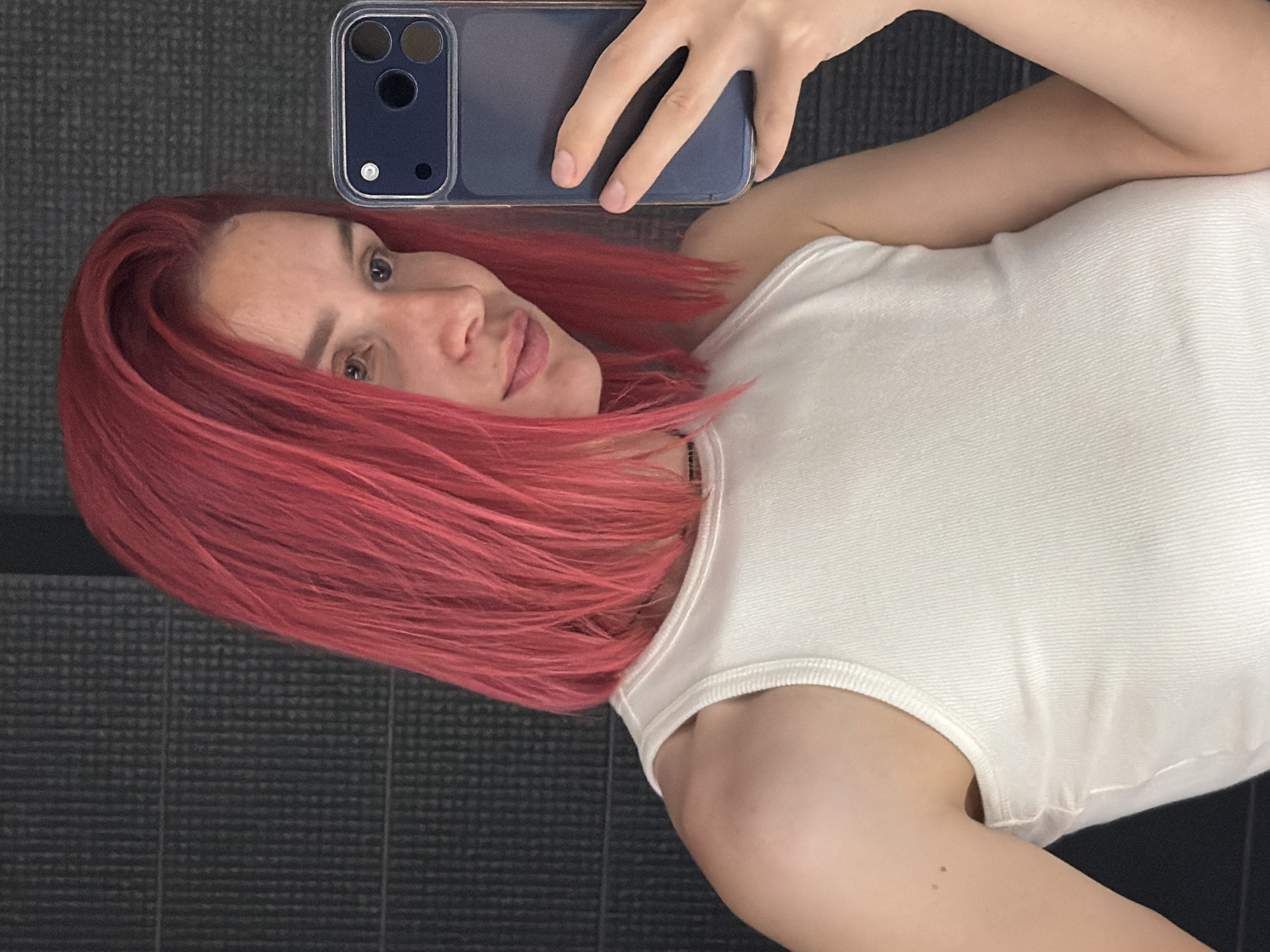 A medium shot captures a woman with vibrant pink hair taking a selfie in what appears to be an indoor setting with a dark, textured wall. She is wearing a white ribbed tank top and is holding a dark blue smartphone in her right hand, angled to capture her reflection. The lighting is somewhat diffused, highlighting the texture of her hair and the ribbed fabric of her top. The background wall features a subtle vertical ribbed pattern, creating a minimalist and somewhat stark environment. No specific location in Chaikovsky, Russia is identifiable from the visual information presented.