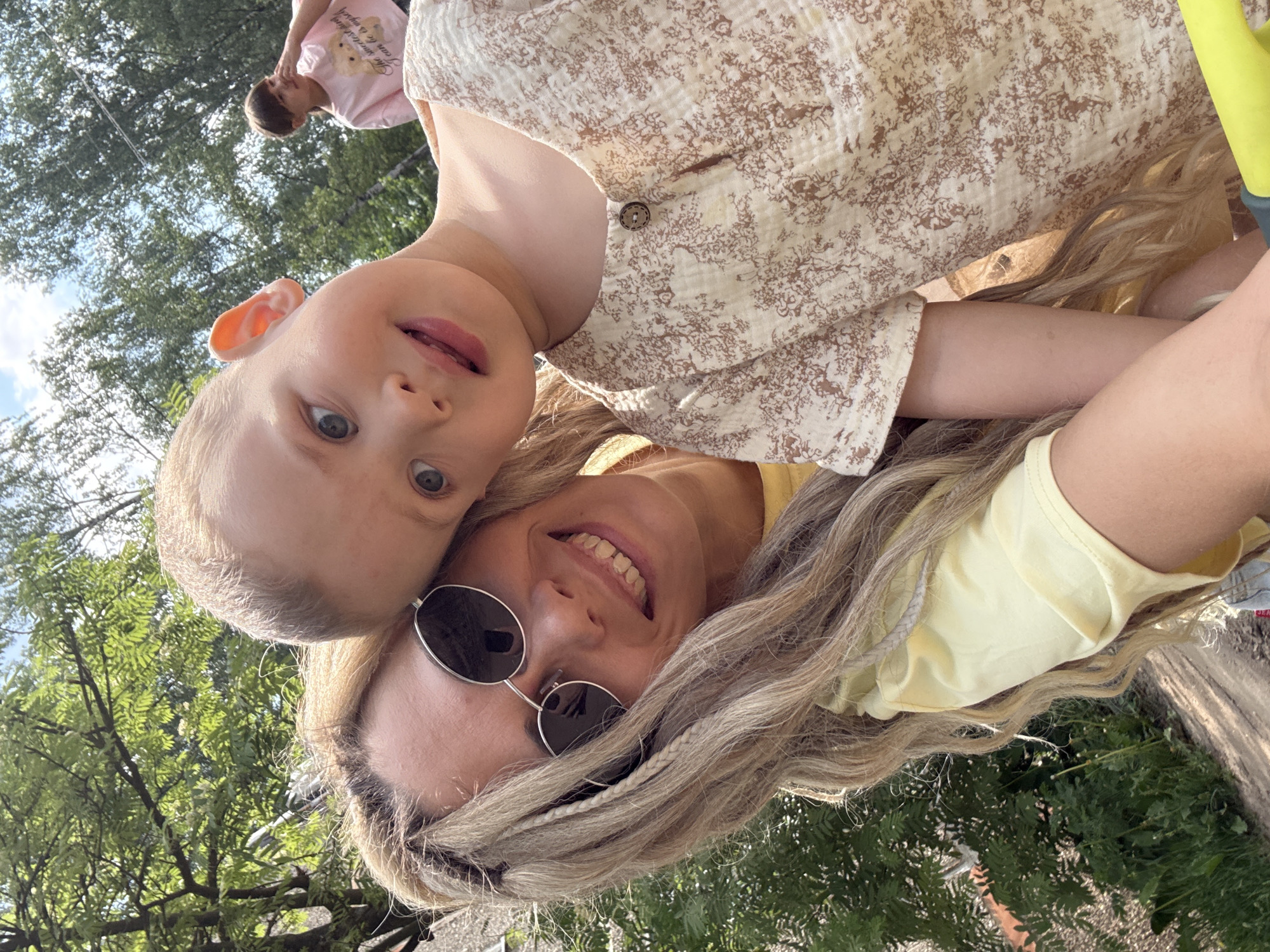 The image shows a woman and a young child in an outdoor setting, likely a park or natural area. The woman is wearing sunglasses and a yellow shirt. Her hair is long and blonde. The child is wearing a floral print shirt. They appear to be taking a selfie. The background includes trees and foliage, suggesting a park or wooded area. There is no information available to identify this image as being in Rusanovo, Russia.