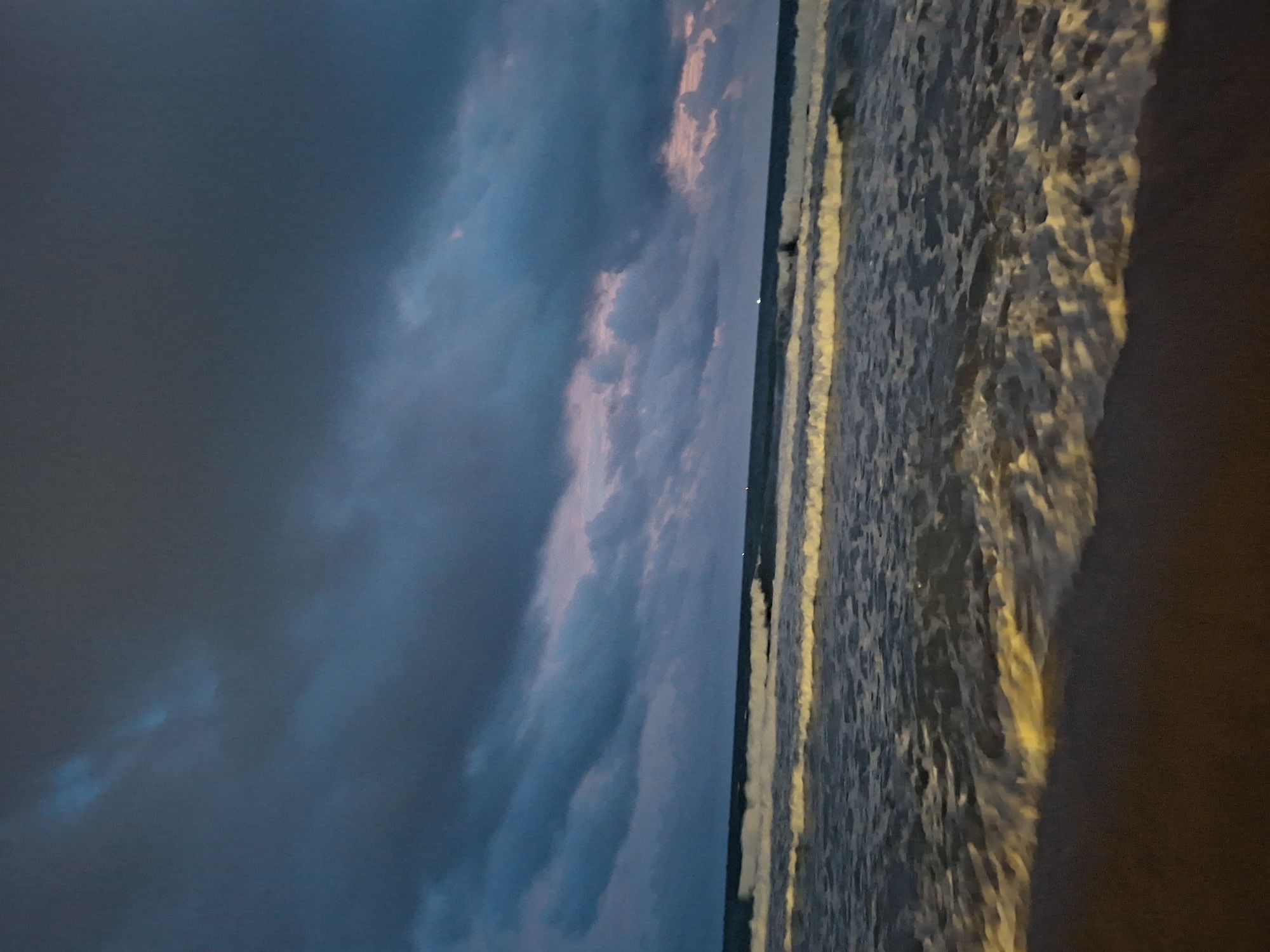 An image depicts the shoreline of a beach, likely in Phu Quoc, Vietnam, at dusk. The sky is overcast with dark, dramatic clouds that transition to hues of pink and blue near the horizon, suggesting the sun has recently set. The sea is active, with several lines of waves breaking on the shore, their crests illuminated by an unseen light source, possibly artificial or the last rays of sunlight. The water appears dark and choppy. The foreground consists of a sandy beach, appearing dark due to the low light conditions. There are no visible people or specific objects in the frame, only the natural elements of sea, sand, and sky. The overall atmosphere is moody and tranquil.