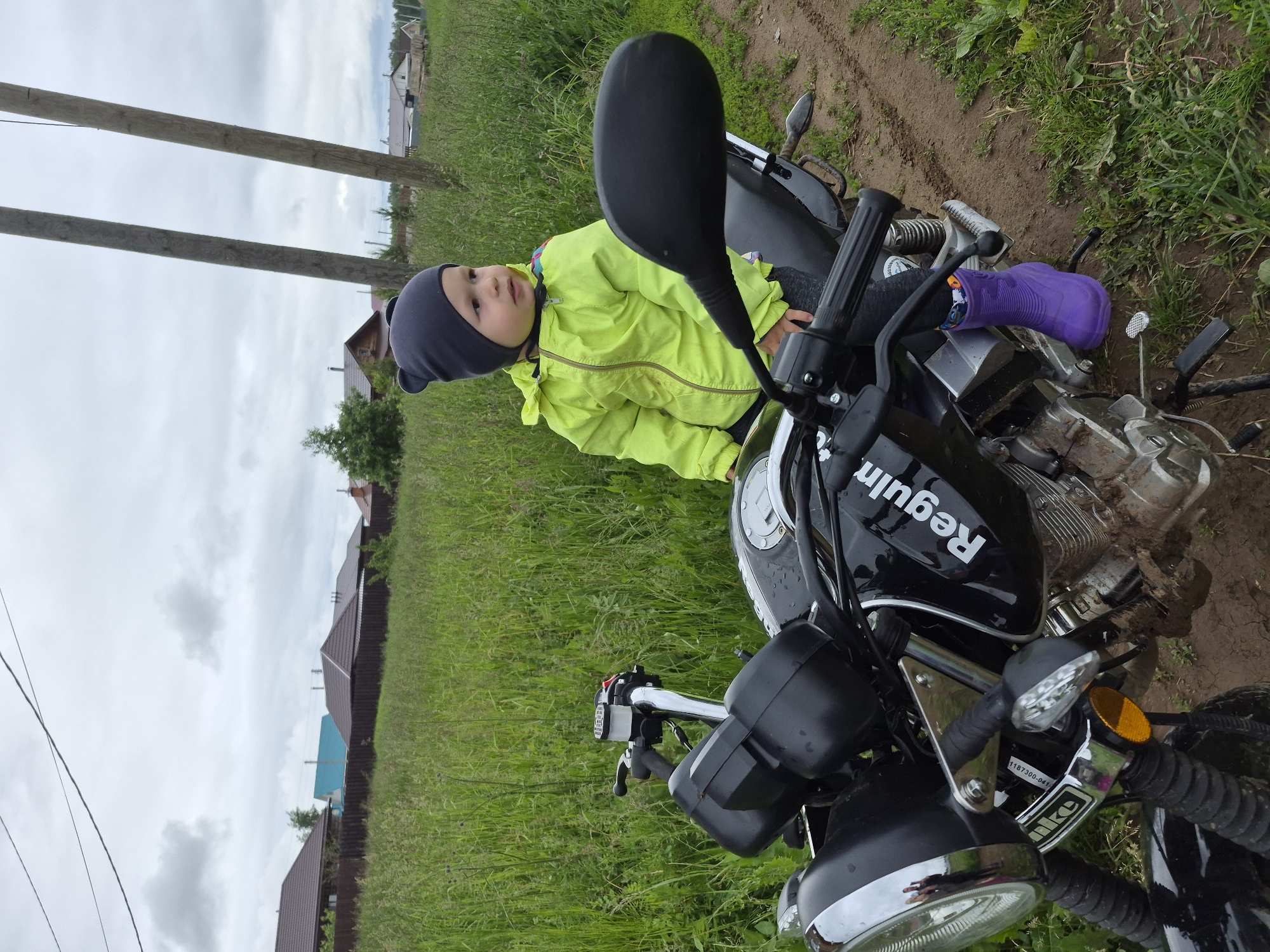 A young child is positioned on a black "Reguln" motorcycle in an outdoor, semi-rural setting under an overcast sky. The child, appearing to be a toddler or preschooler, wears a neon yellow jacket, dark pants, a dark grey knit beanie with bear ears, and a single vibrant purple rubber boot on their right foot. They are standing on the left side of the motorcycle, with their left leg over the seat and their torso leaning against the fuel tank. The child is looking towards the right with a neutral expression.
The motorcycle is a black model with "Reguln" written in white on the fuel tank. It exhibits mud splatters on the engine block and lower frame, indicating recent off-road use. Visible features include a chrome headlight bezel, clear front turn signals with an orange reflector below the left one, black handlebars with a visible front brake fluid reservoir on the right, and a black rearview mirror on the right side. The motorcycle is stationary on what appears to be a dirt path, with tall green grass and weeds surrounding it and extending into the background.
In the mid-ground, a dark brown wooden fence separates the grassy area from several houses with brown and one with a blue roof. Two weathered wooden utility poles are prominent on the right side of the image. Power lines are faintly visible against the light grey, cloudy sky. The ground immediately around the motorcycle is a mix of dirt and sparse vegetation. The location is in the city of Unknown, Unknown.