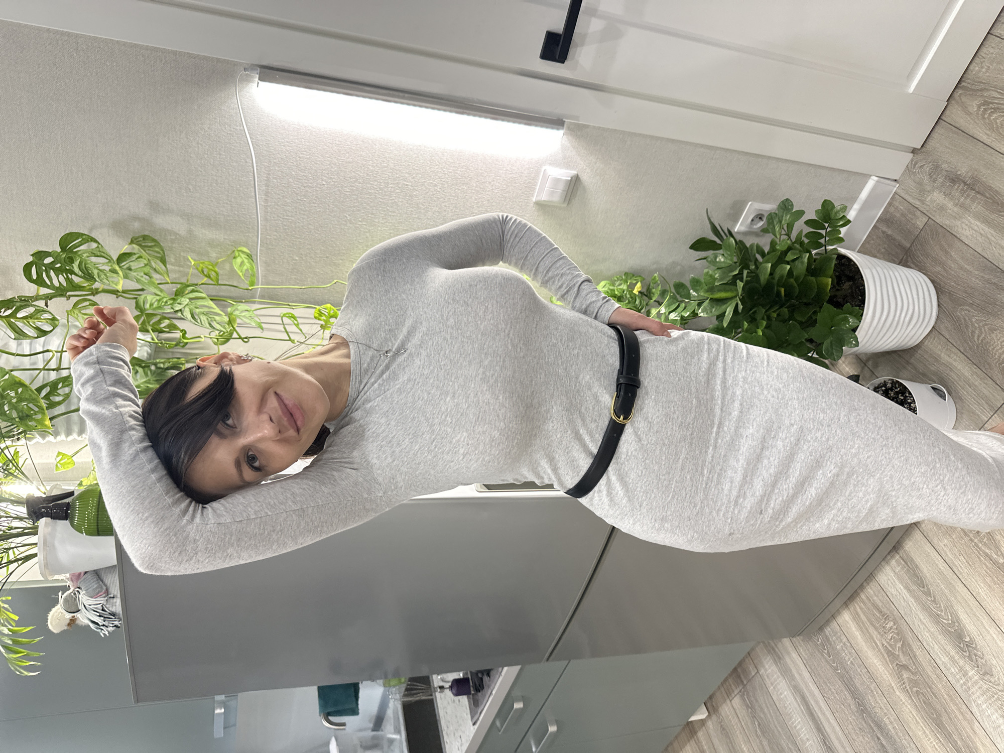 A woman wearing a gray long-sleeved dress and a black belt is indoors, leaning on a gray countertop. Her left arm is raised and resting on the edge of the countertop, while her right hand is on her hip. To her left are two potted plants, one with vines cascading down the side. Behind her, there is another potted plant and a white door. The flooring is a light wood laminate. Above, there is a strip light attached to the wall.