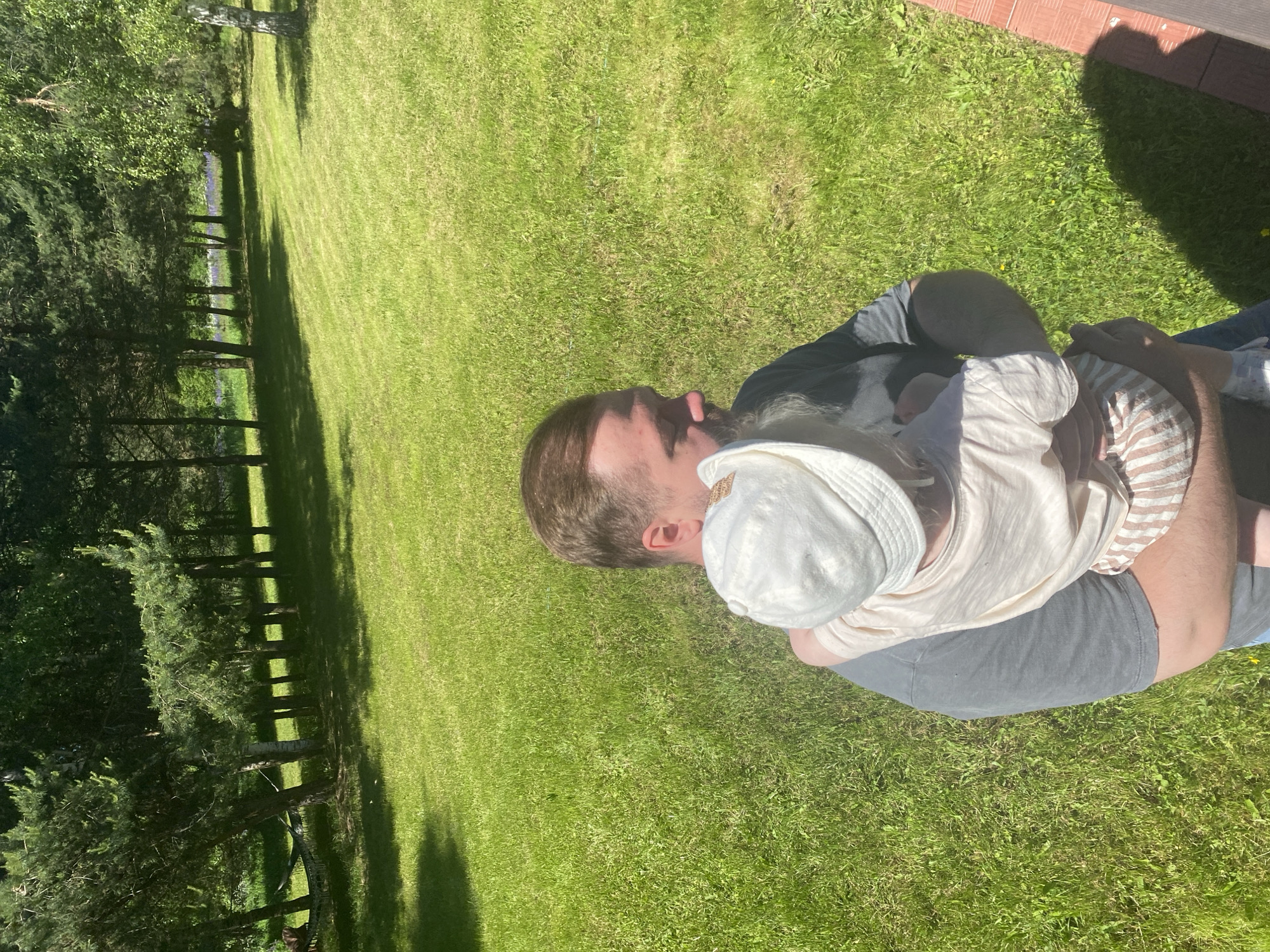 A bearded man, wearing a dark grey t-shirt and blue jeans, holds an infant or toddler outdoors in Копнино, Russia. The man's head is turned to his left, away from the direct camera view. The child, with their back to the camera, is wearing a light-colored bucket hat, a light-colored t-shirt, and striped shorts, resting against the man's chest. The environment consists of a vast, mown green lawn under direct sunlight, casting distinct shadows. A line of pine and deciduous trees forms the background, with a hammock visible hanging between trees on the left. A small section of a reddish-brown tiled or wooden structure is visible in the lower right foreground, partially obscured by a human shadow.